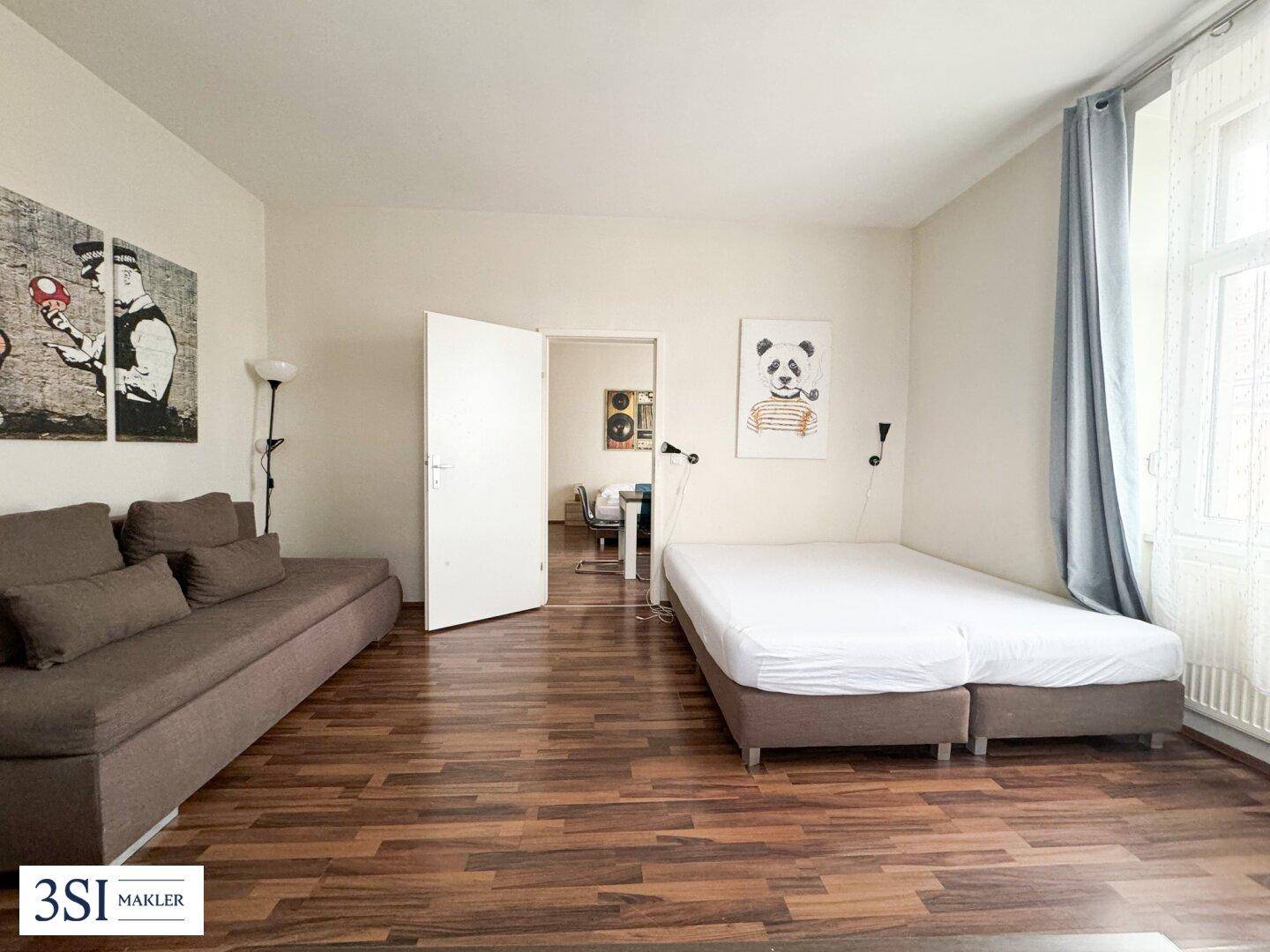 Gemütlicher Wohnbereich mit Sofa, Bett und Holzboden, durch eine Tür mit einem weiteren Raum verbunden.