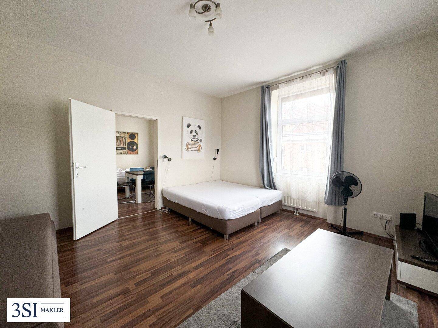 Wohn- und Schlafbereich mit Sofa, Bett und Blick in einen angrenzenden Raum mit Holzboden.