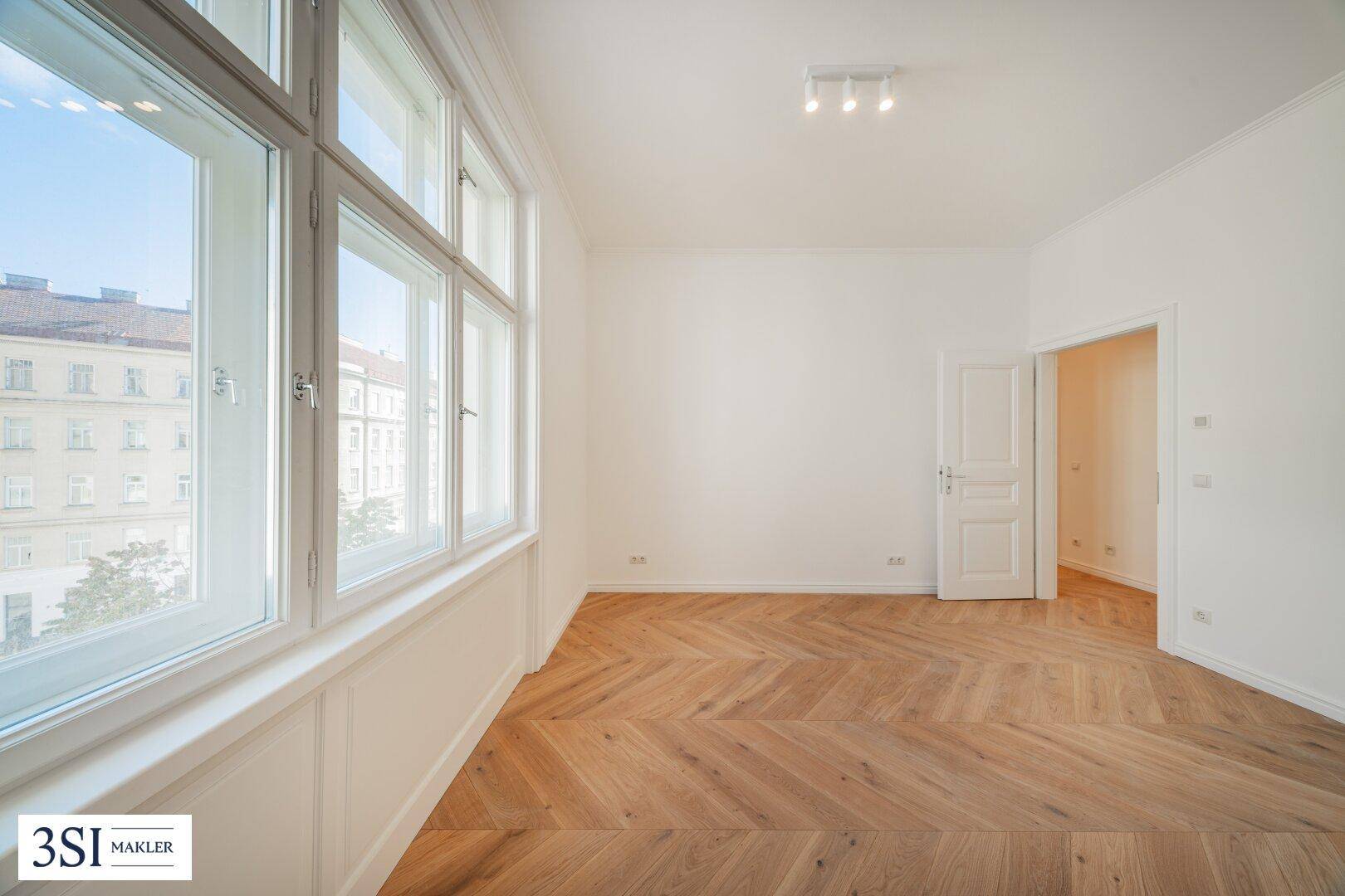 Heller, leerer Raum mit Fischgrätparkett und großen Fenstern mit Stadtblick.