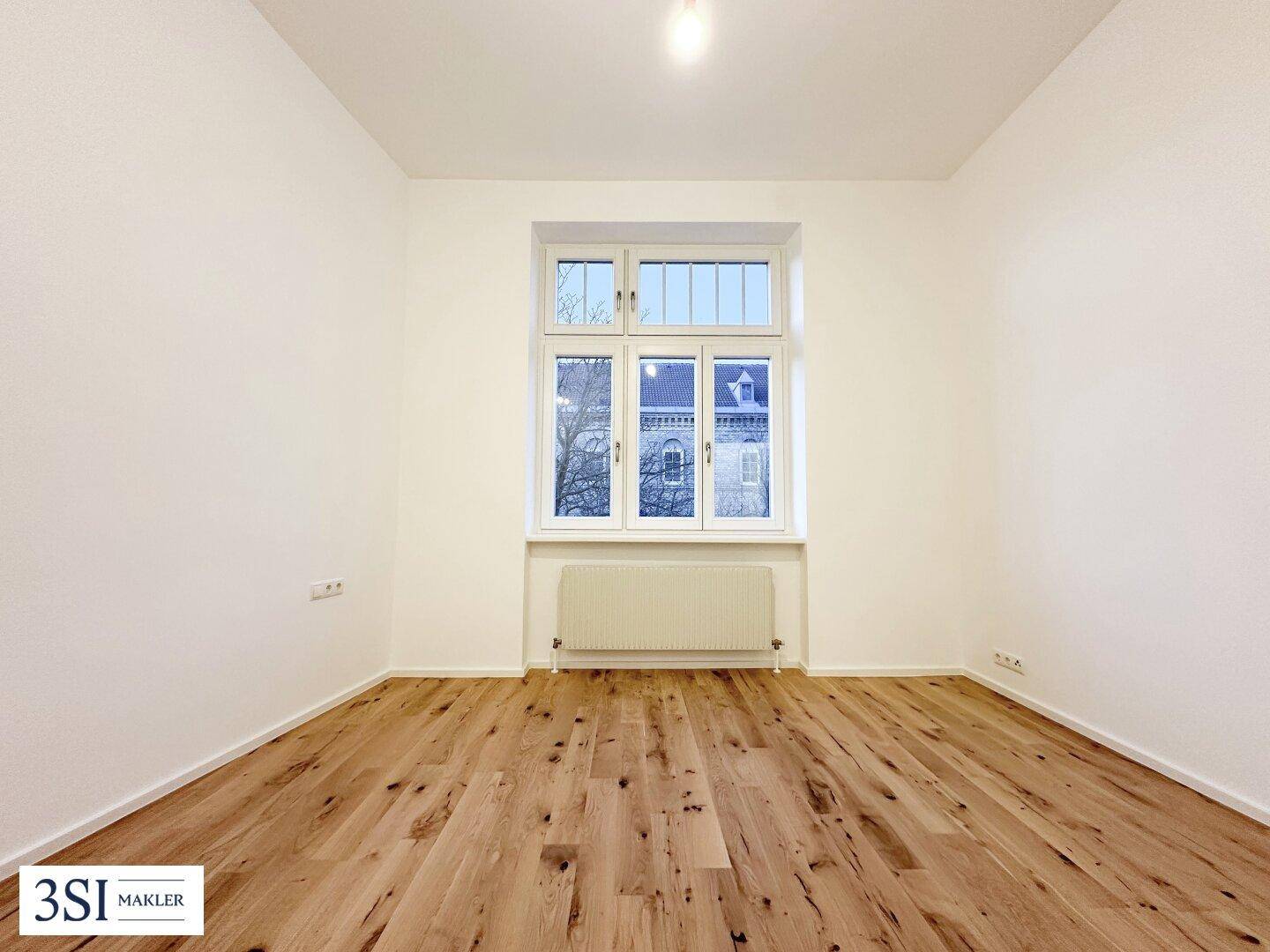 Freundliches Zimmer mit großem Fenster, Holzboden und Heizkörper unter dem Fenster.