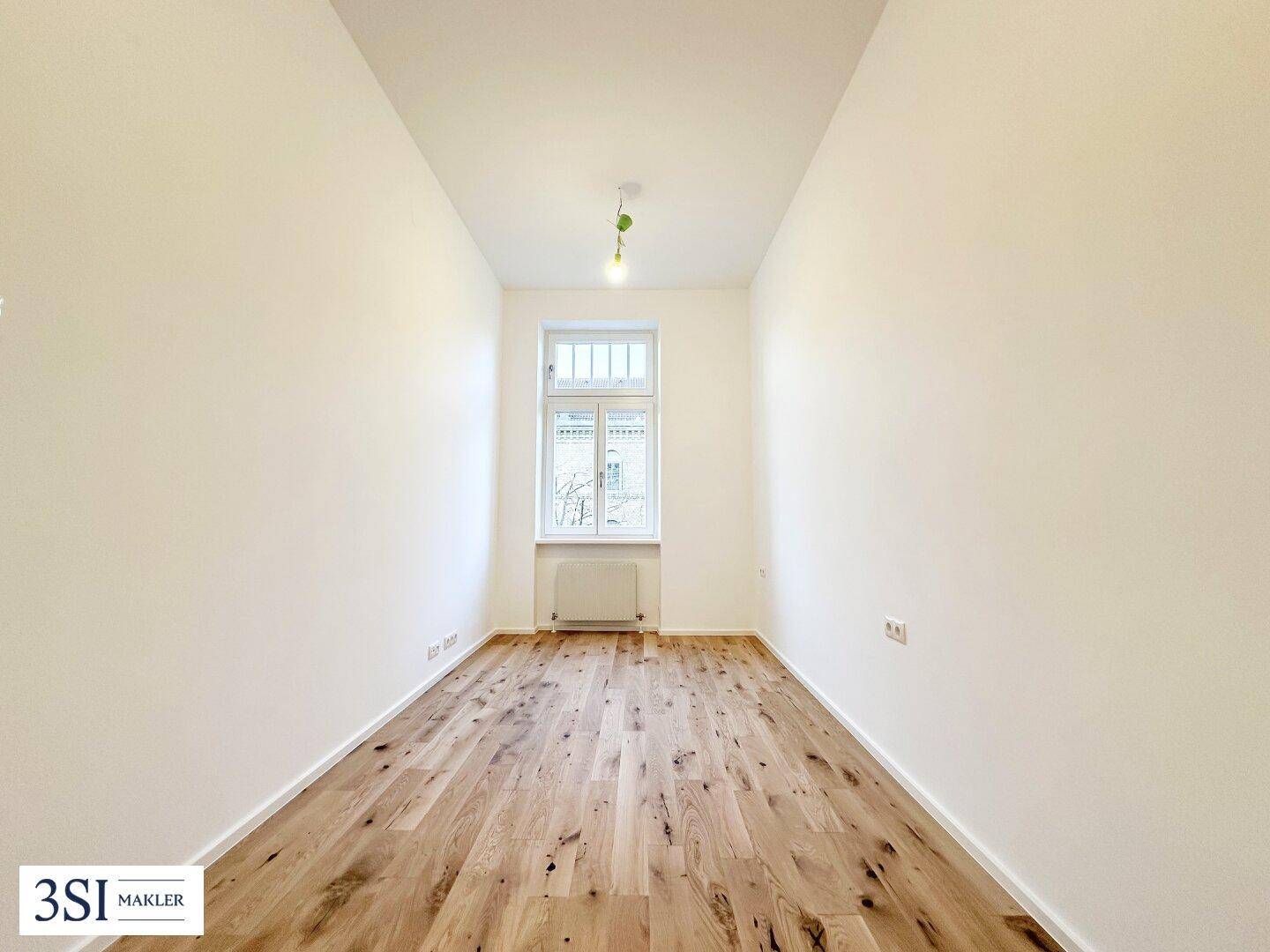 Schmaler Raum mit Holzboden und einem Fenster, das viel Tageslicht hereinlässt.