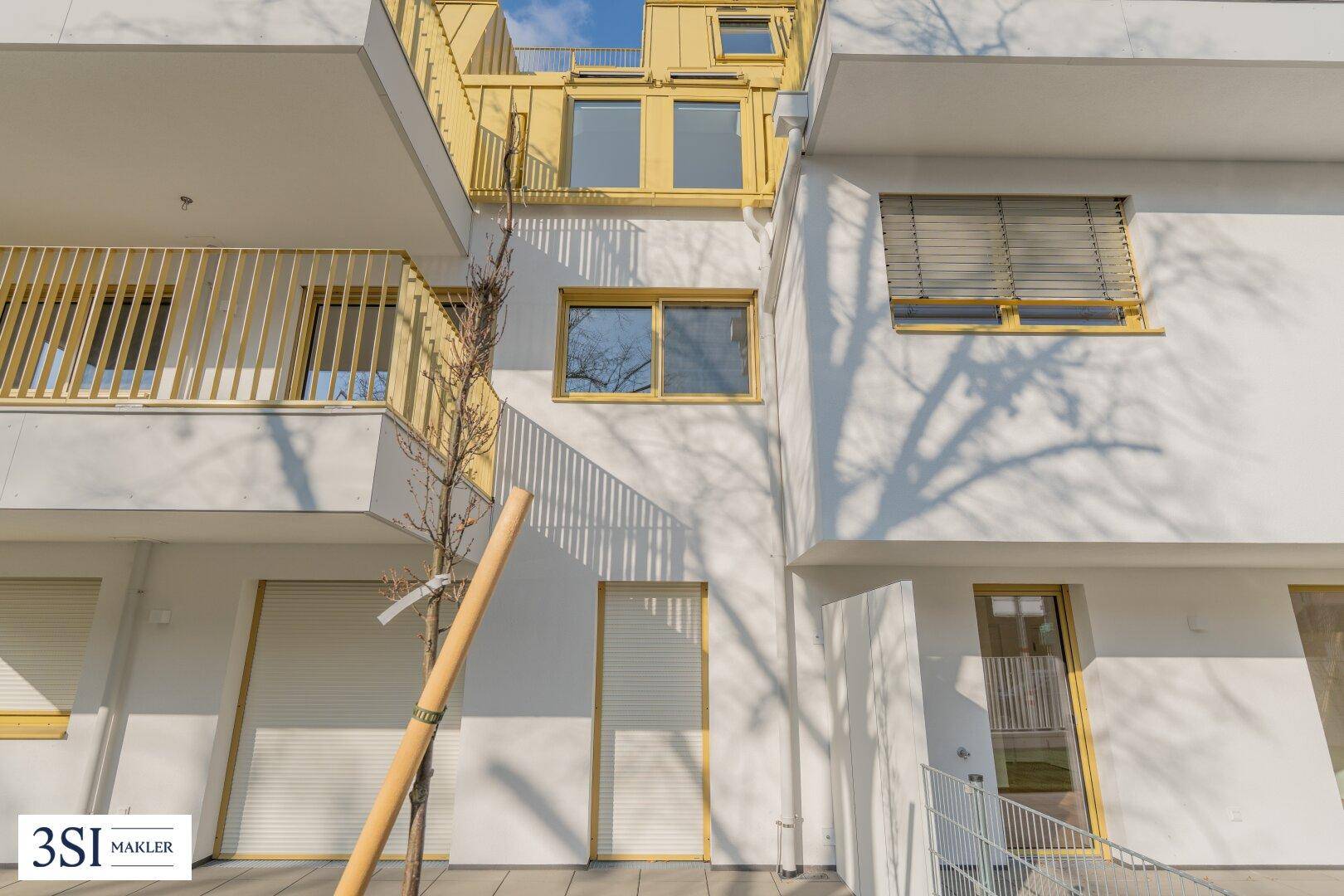 Außenansicht eines modernen Gebäudes mit Terrasse, Balkon und geschlossenen Rollläden bei Sonnenschein.
