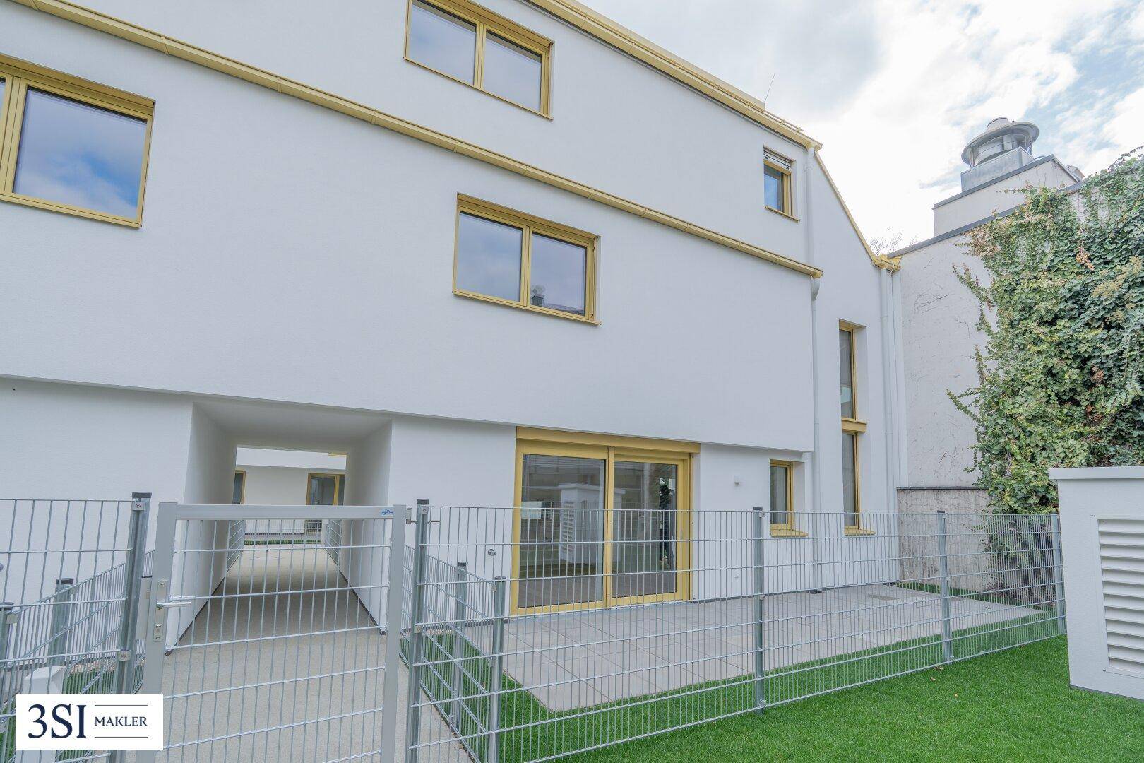 Hinterer Bereich des Gebäudes mit kleiner Rasenfläche, Terrasse und Zugang durch eine Schiebetür.