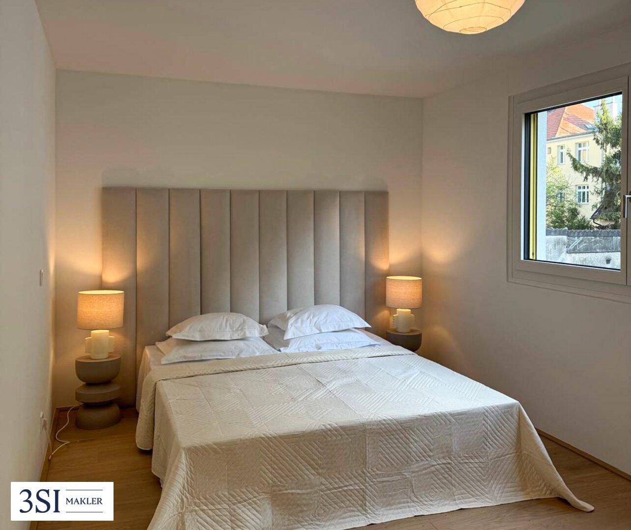 Ruhiges Schlafzimmer mit beigem Kopfteil, Holzboden und einem Fenster mit Blick nach draußen.
