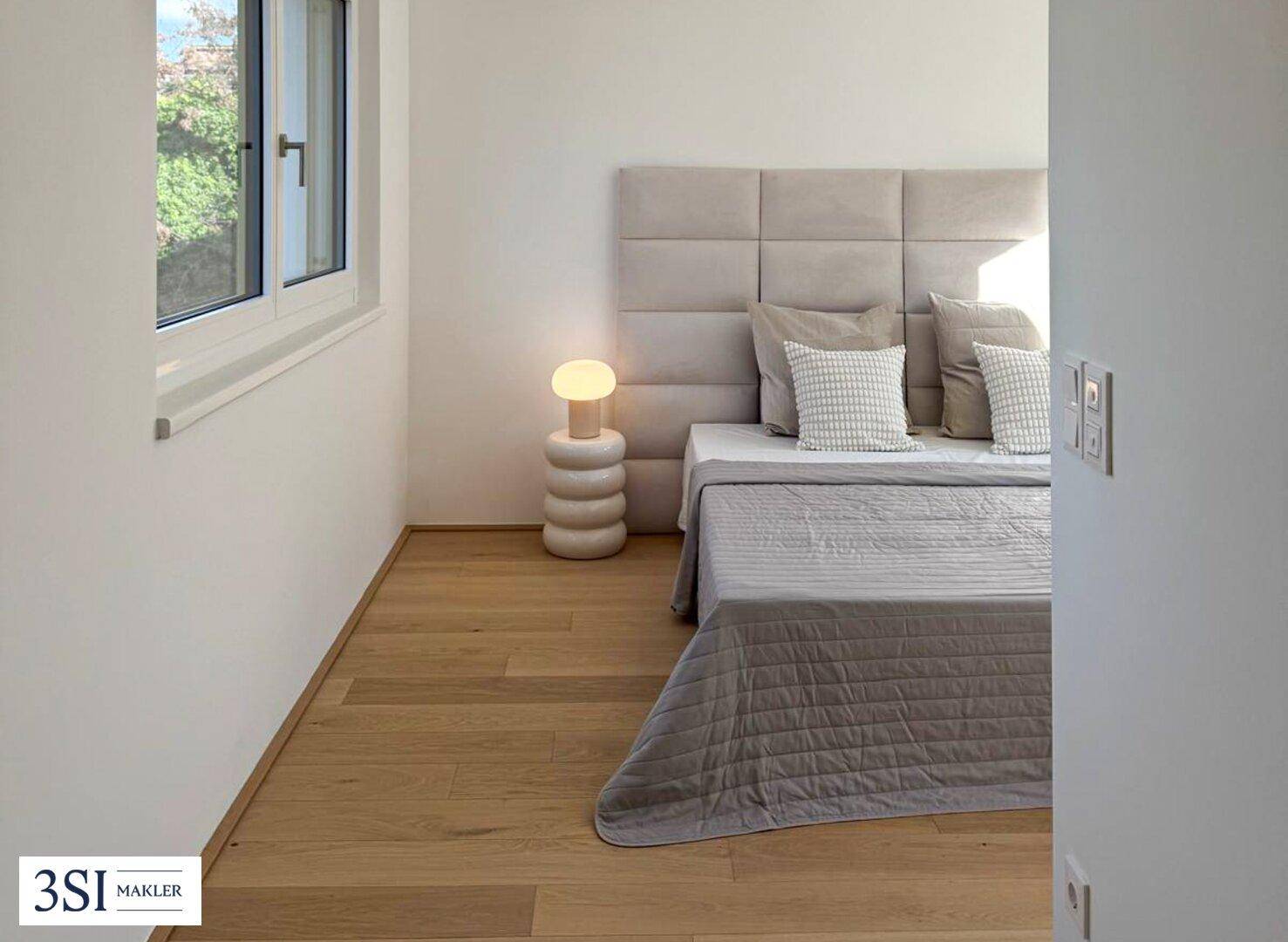 Helles Schlafzimmer mit Holzboden, gepolstertem Kopfteil und Blick ins Grüne.