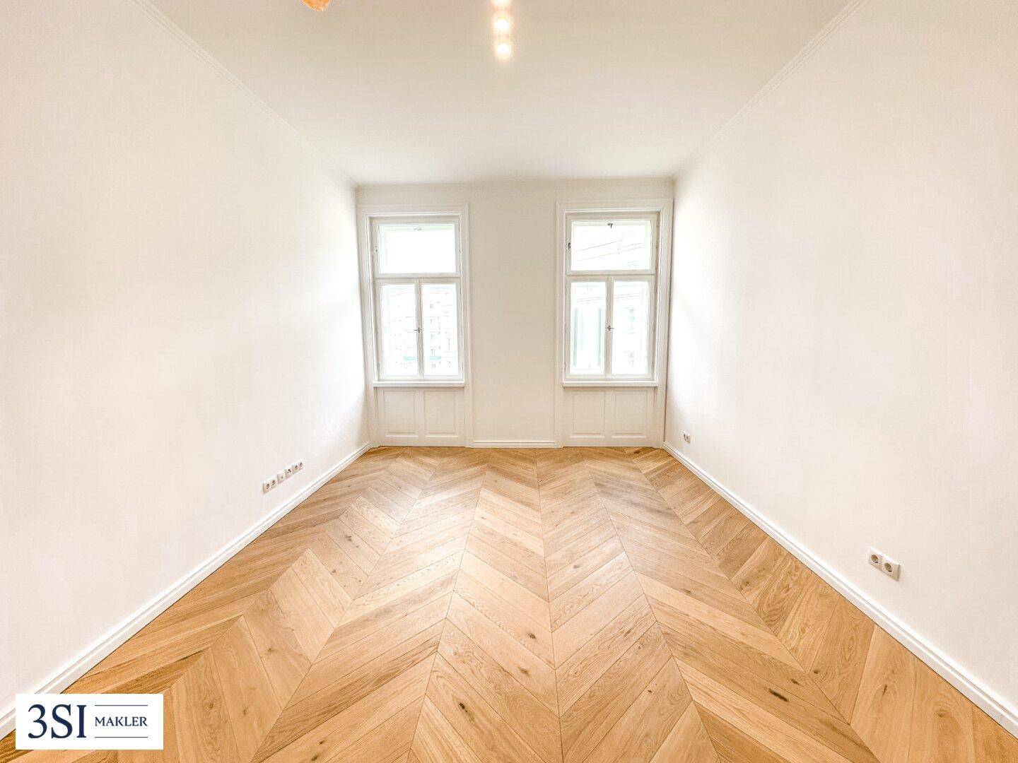 Helles, leeres Zimmer mit Fischgrätparkett und zwei Fenstern, ideal als Schlaf- oder Arbeitszimmer.