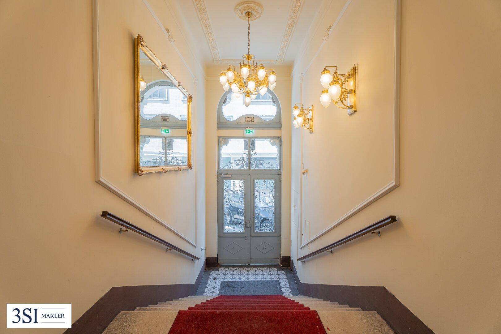 Repräsentatives Treppenhaus mit rotem Teppich, Stuckdecke und klassischer Beleuchtung.