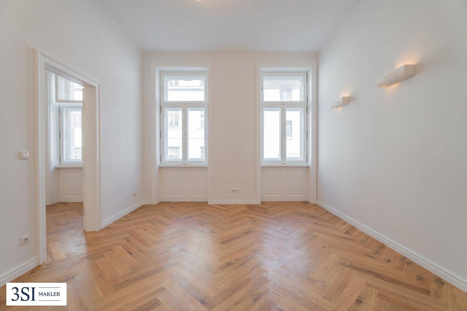 Geräumiges Zimmer mit Fischgrätparkett, hohen Decken und viel Tageslicht durch große Fenster.