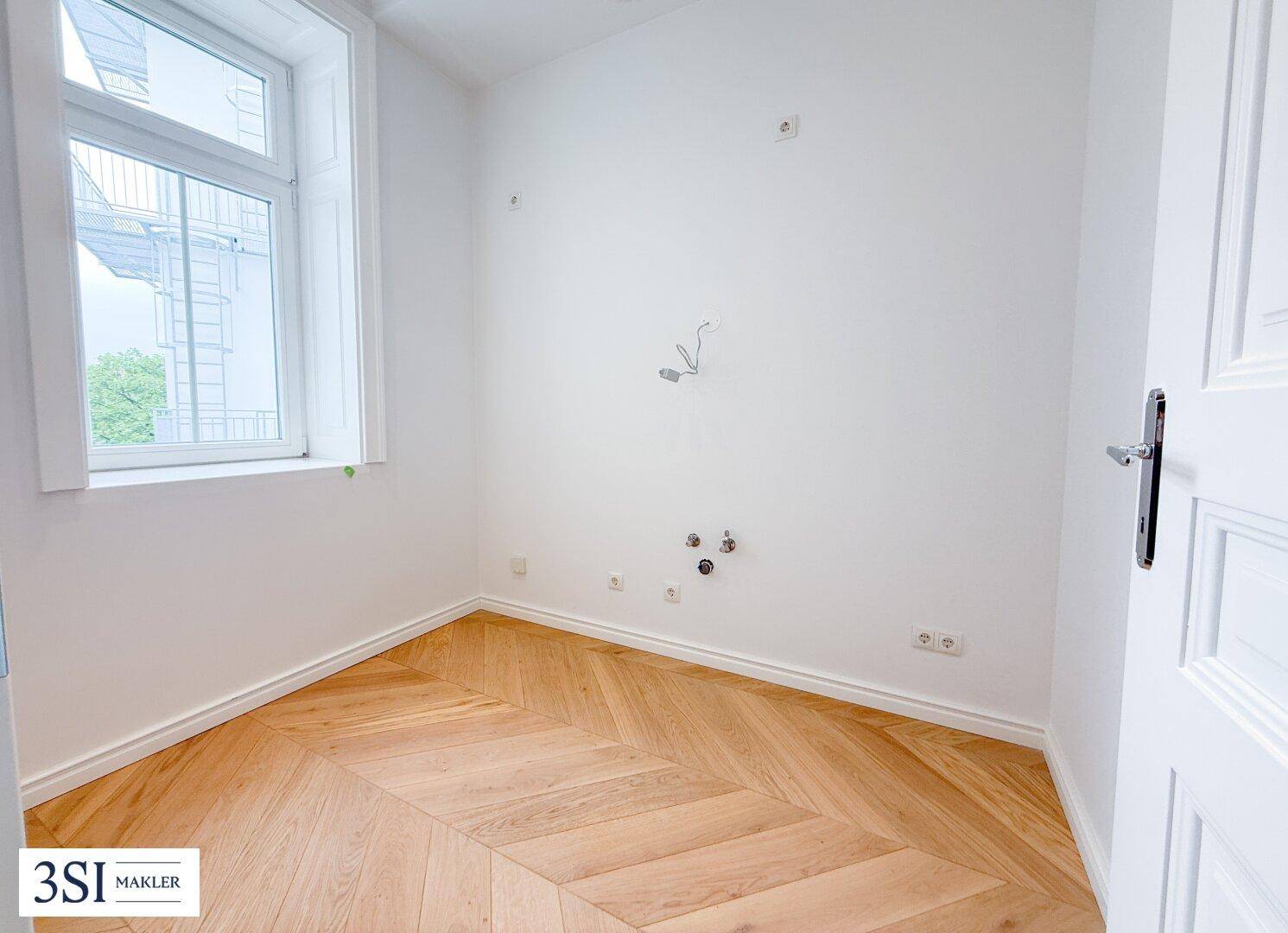 Leerer Raum mit Anschlüssen für eine Küche und elegantem Fischgrätparkett.