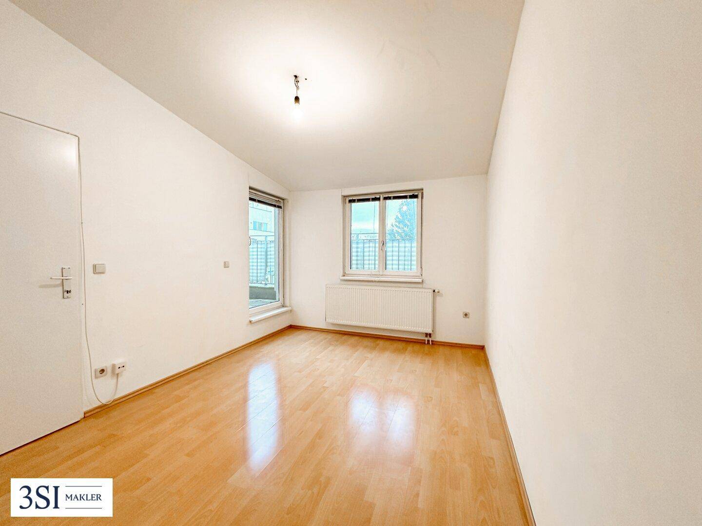 Leeres Zimmer mit Holzboden, hellen Wänden und einem französischen Balkon.
