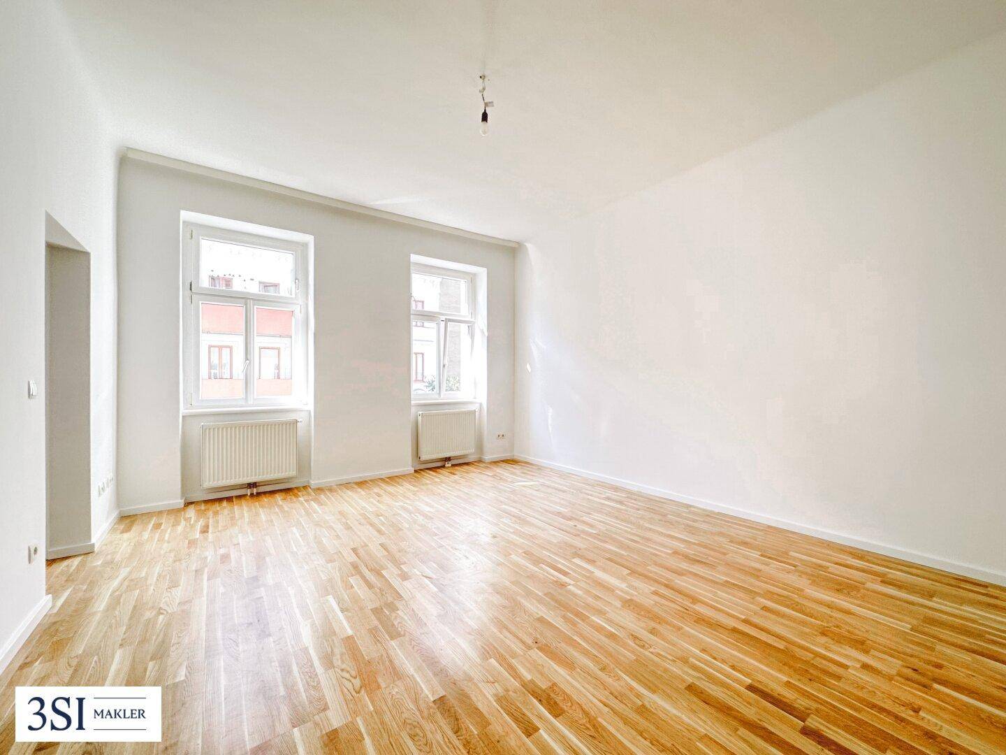 Geräumiges, leeres Wohnzimmer mit Holzboden und zwei großen Fenstern, bereit zur individuellen Gestaltung.