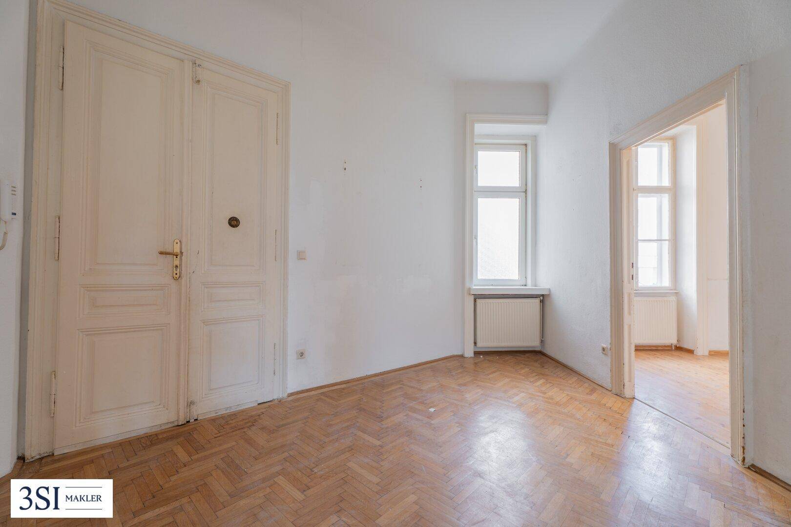 Geräumiges Zimmer mit Fischgrätparkett, hohen Türen und Blick in einen weiteren Raum.