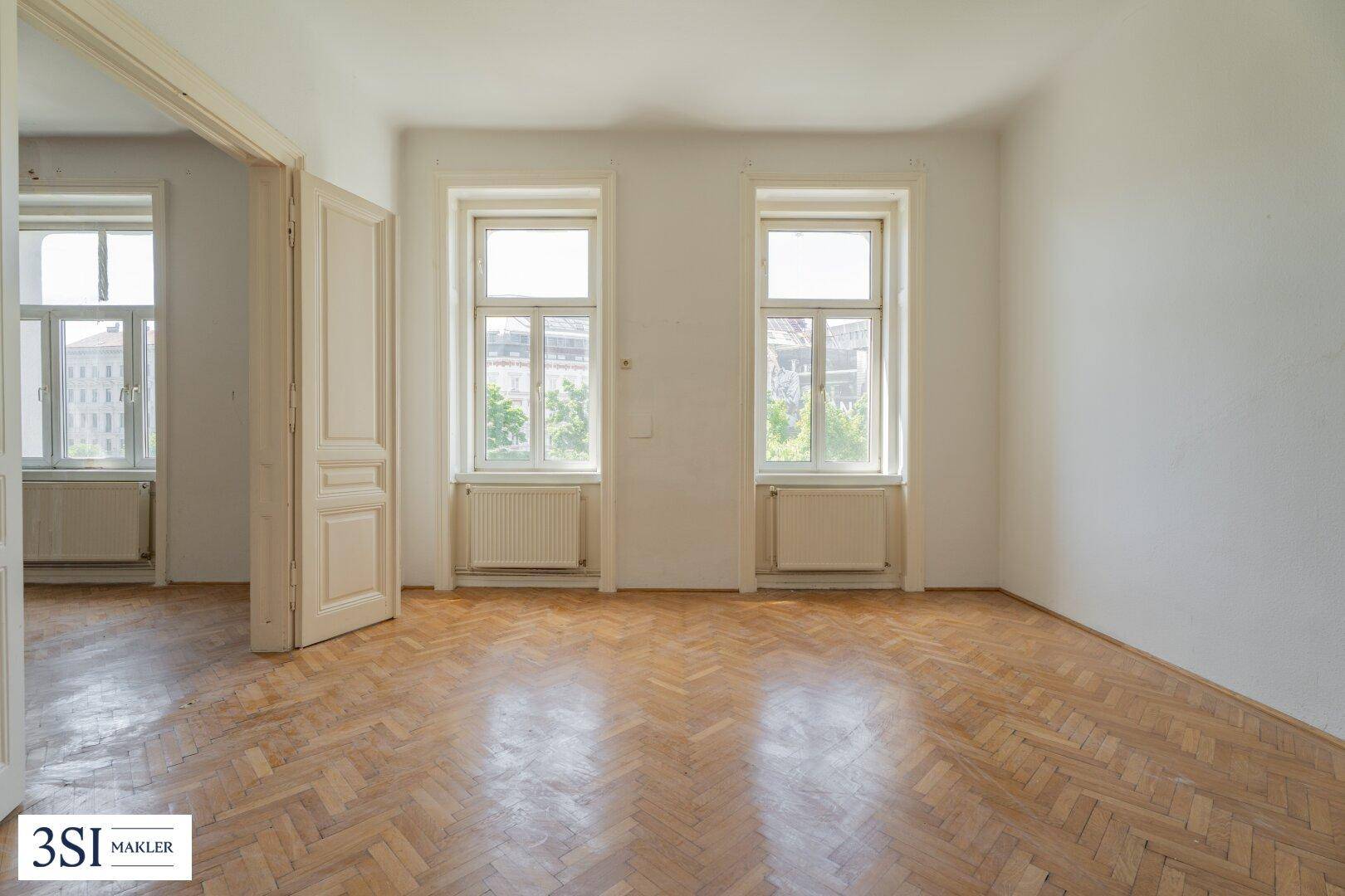 Helles Zimmer mit zwei Fenstern, Fischgrätparkett und Zugang zu einem weiteren Raum.