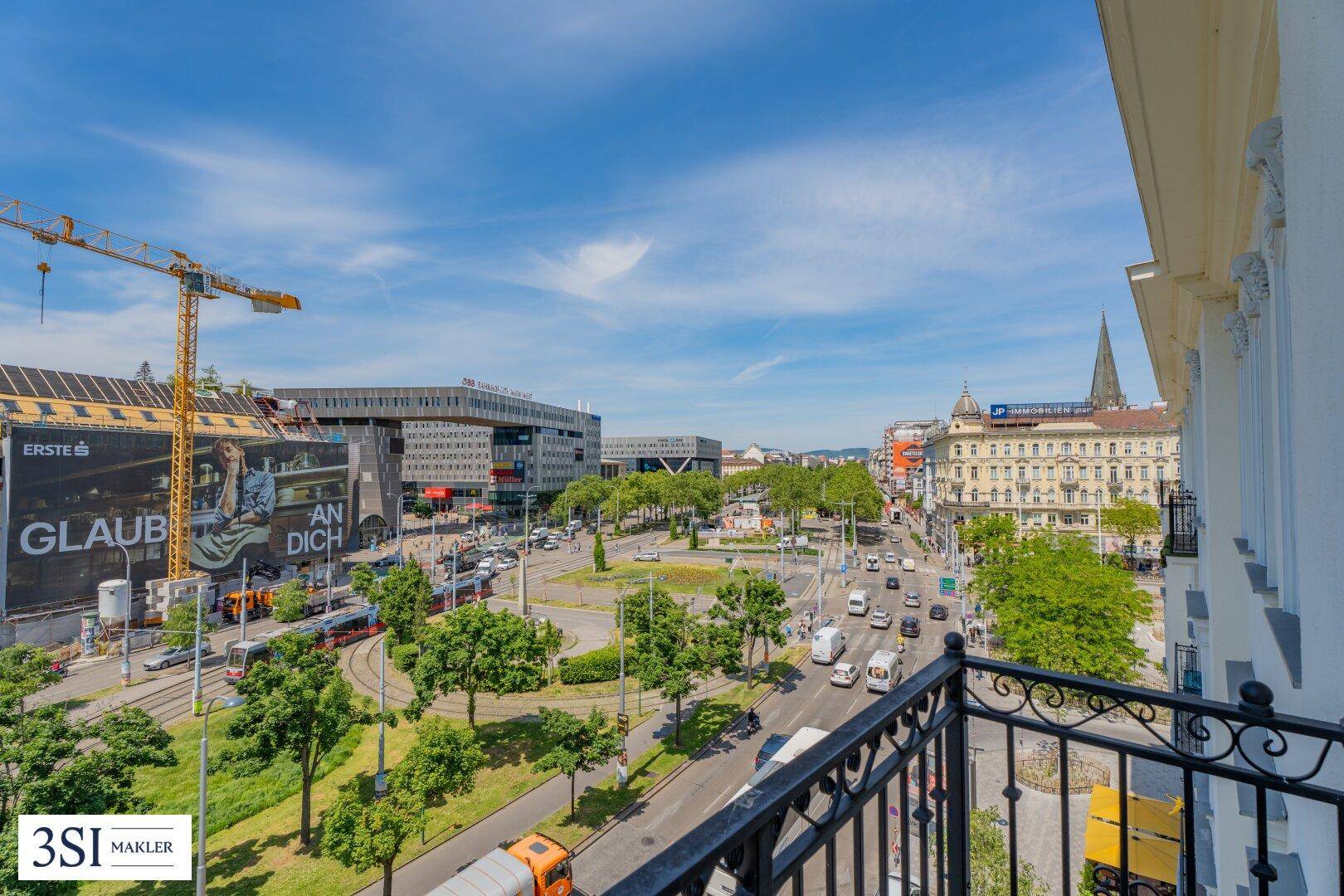 Blick vom Balkon auf eine belebte Stadtkreuzung mit Gebäuden und viel Grün.
