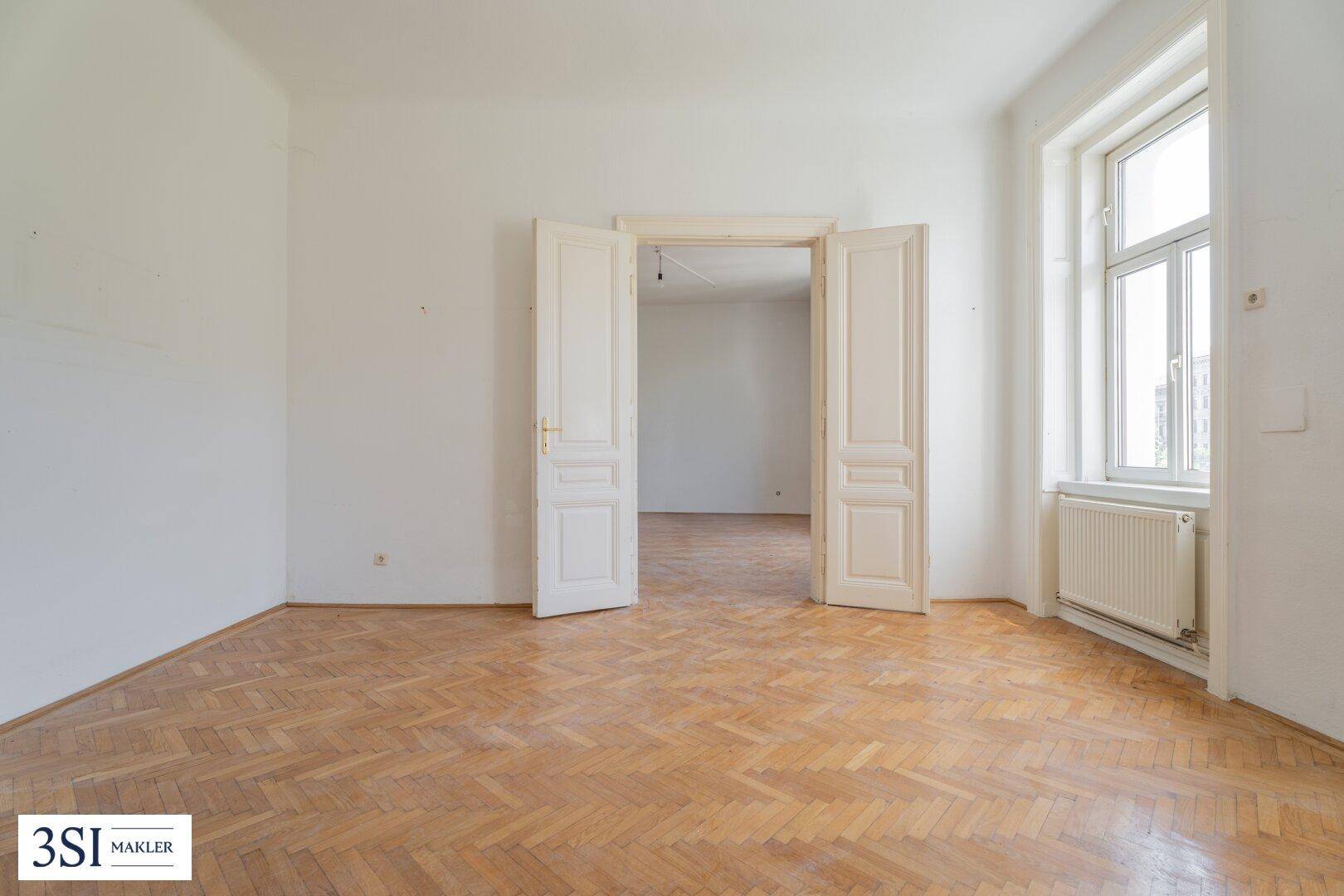 Großes Zimmer mit Fischgrätparkett und einer breiten Doppeltür zu einem angrenzenden Raum.