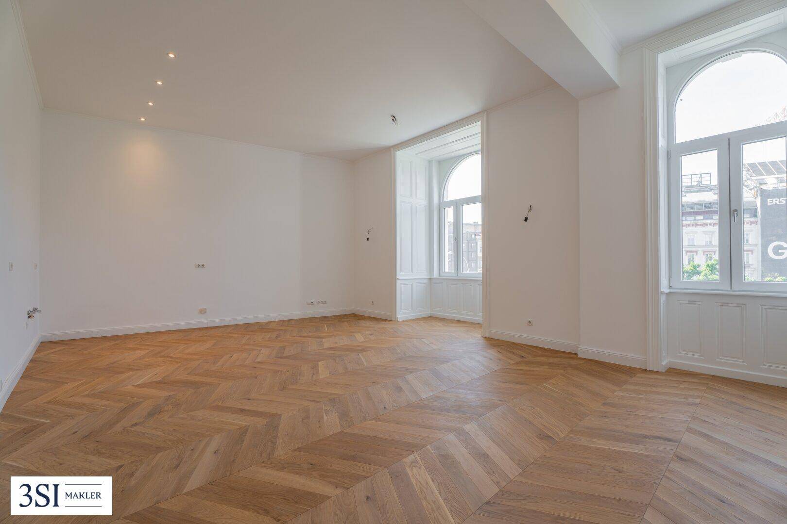 Geräumiges Zimmer mit Fischgrätparkett, hohen Decken und großen Bogenfenstern.