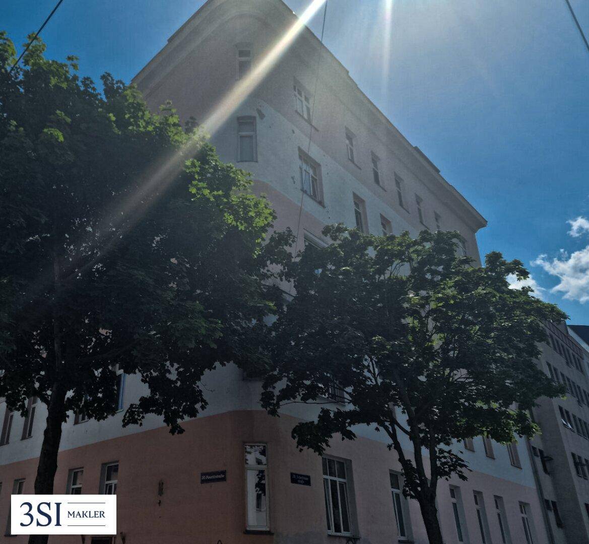 Helle Gebäudefassade mit Bäumen im Vordergrund unter blauem Himmel, beleuchtet von der Sonne.