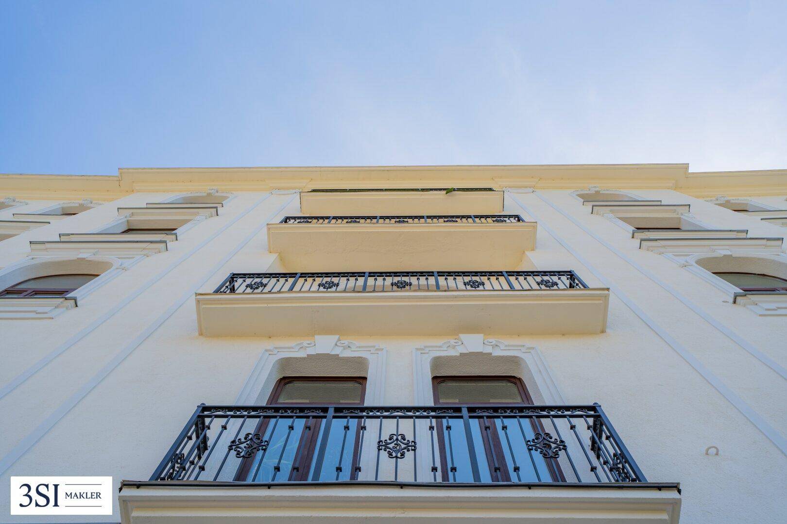 Die helle Fassade des Gebäudes mit mehreren Balkonen und kunstvollen Geländern unter blauem Himmel.