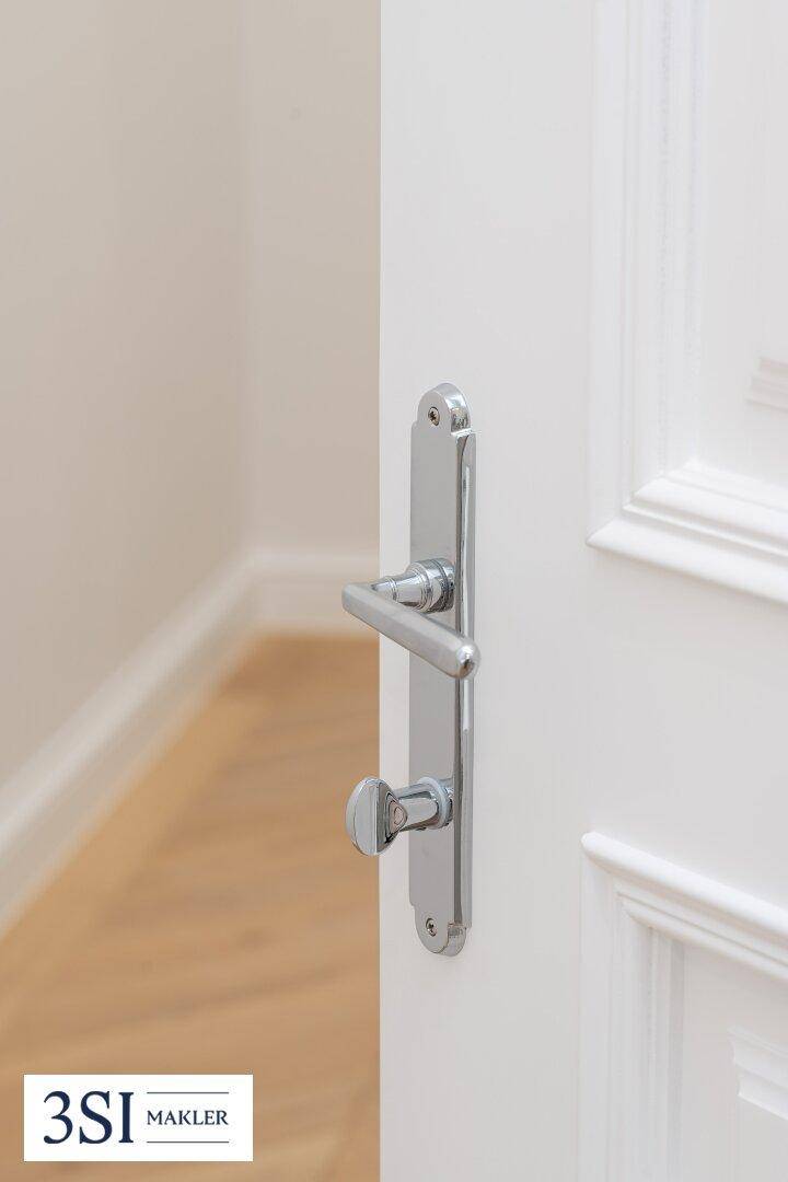 Detailaufnahme einer weißen Innentür mit elegantem Chromgriff, die einen Raum mit hellem Holzboden und weißer Wand freigibt.