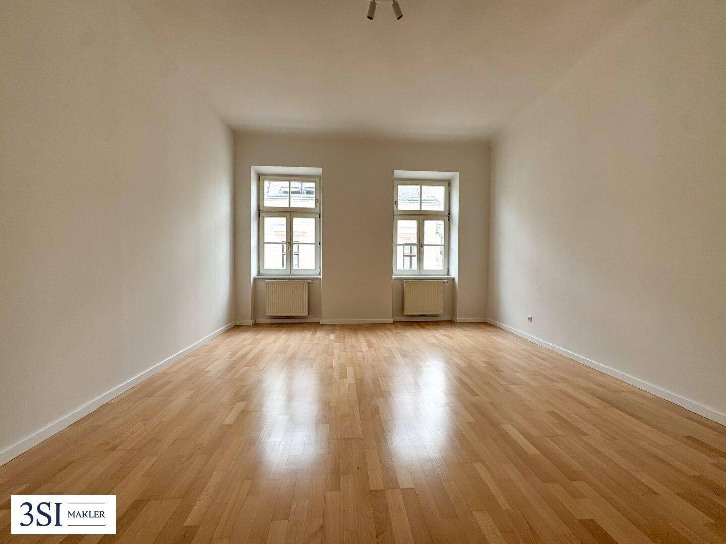Leeres Zimmer mit hellem Parkettboden, beigen Wänden und zwei Fenstern, die für natürliche Belichtung sorgen.