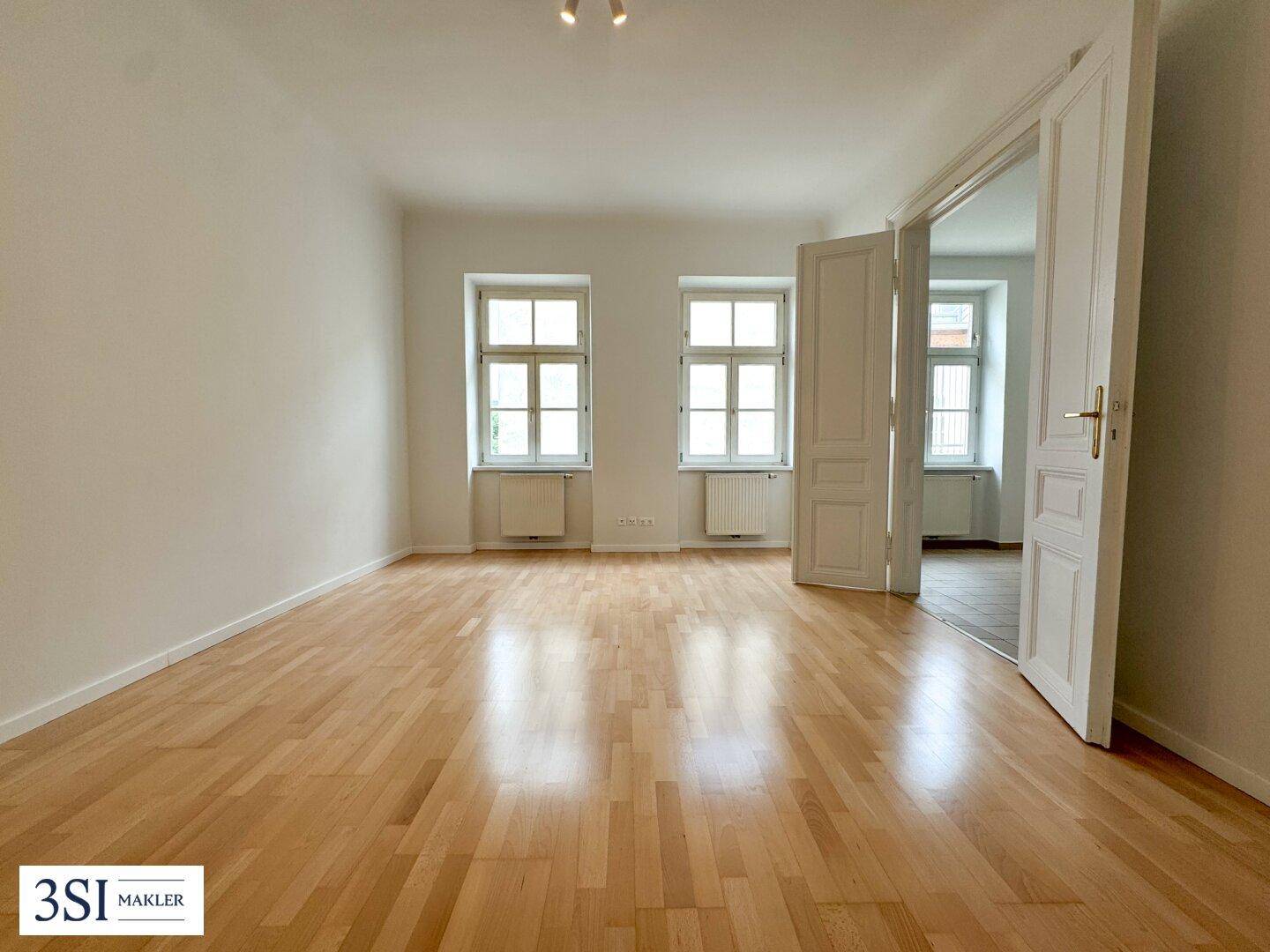 Helles, leeres Zimmer mit Parkettboden, weißen Wänden und hohen Fenstern, das zu einem weiteren Raum führt.