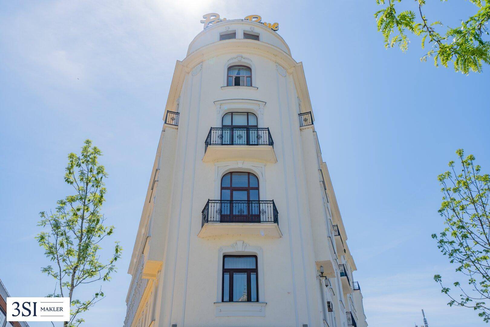 Abgerundete Eckfassade des Gebäudes mit mehreren Balkonen und dekorativen Elementen unter blauem Himmel.
