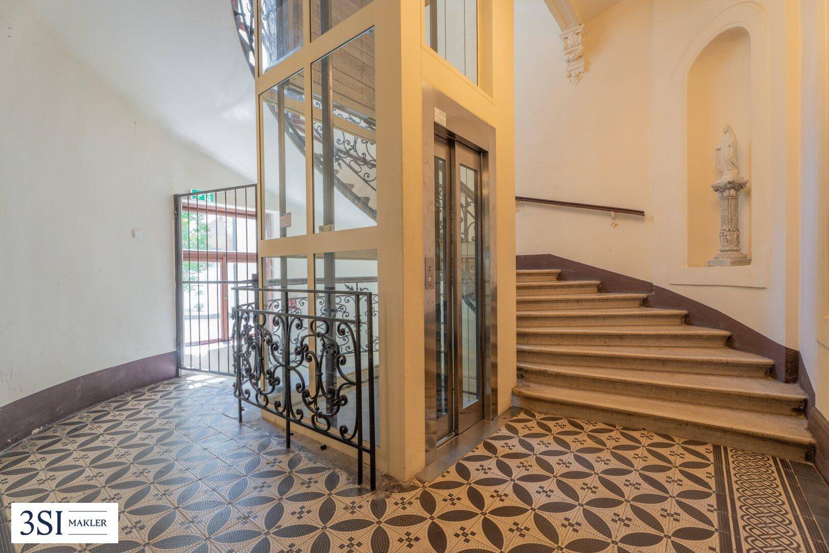 Eleganter Eingangsbereich mit Glasaufzug, geschwungener Treppe und gemusterten Bodenfliesen.