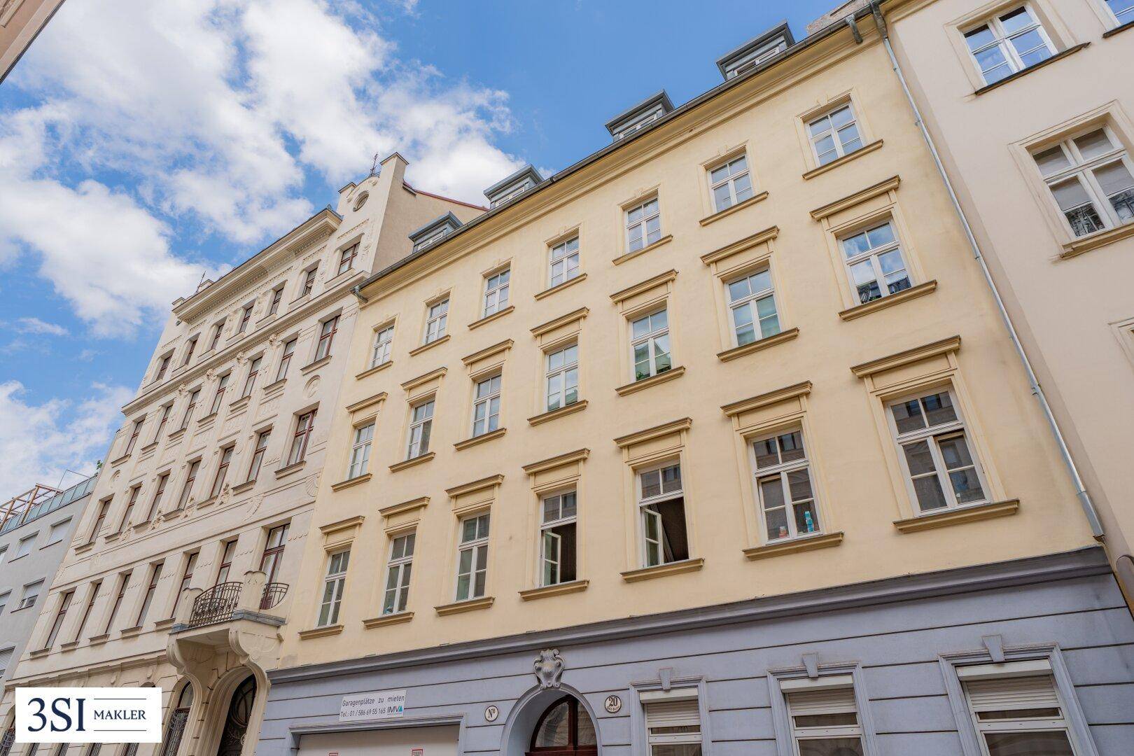 Gesamtansicht der beigen Altbaufassade mit vielen Fenstern und angrenzenden Gebäuden in einer städtischen Umgebung.