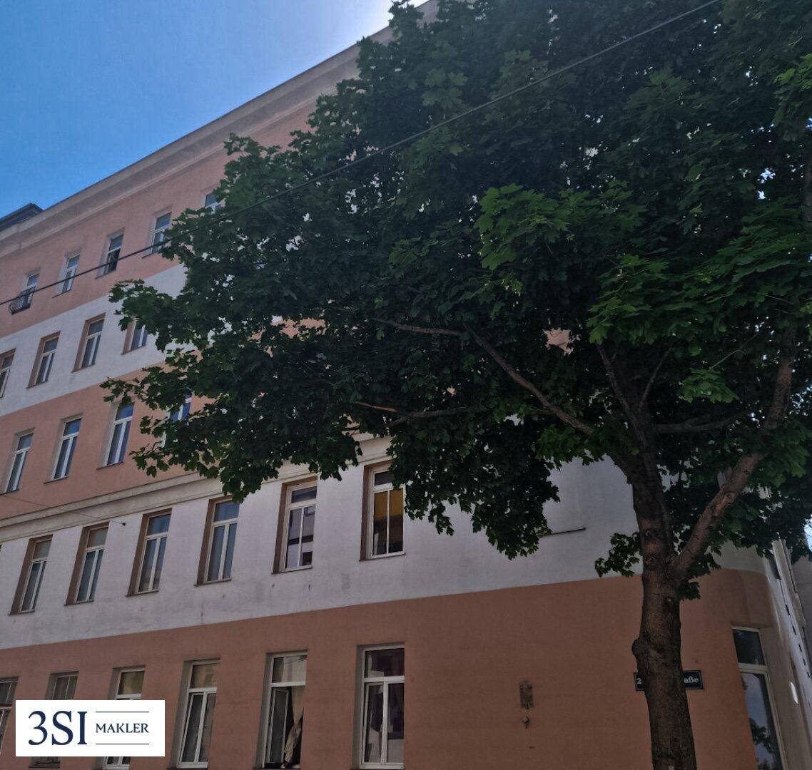 Fassade eines Eckgebäudes mit hohen Bäumen im Vordergrund unter blauem Himmel.