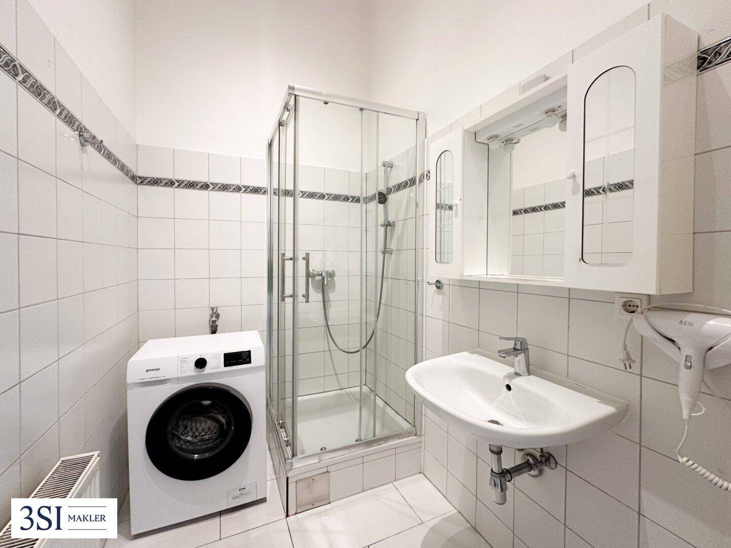 Funktionales Badezimmer mit Duschkabine, Waschmaschine und weißen Fliesen an Wänden und Boden.