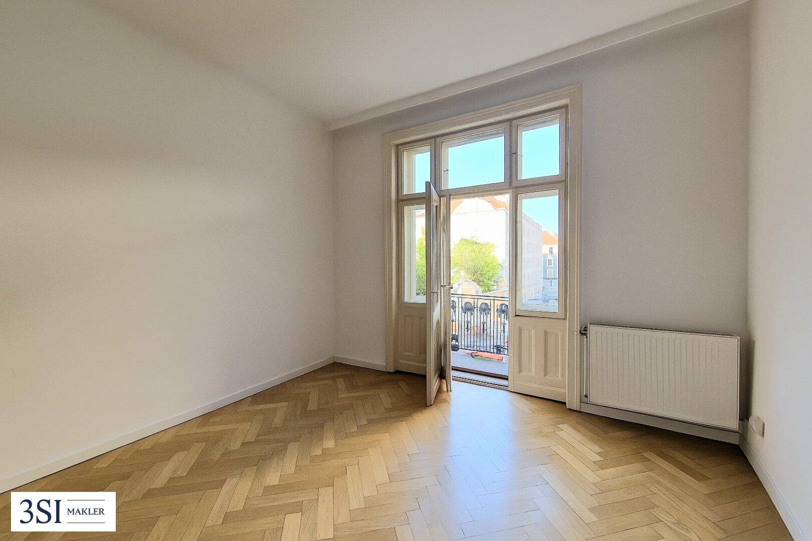 Lichtdurchfluteter Raum mit Fischgrätparkett und Zugang zu einem französischen Balkon.