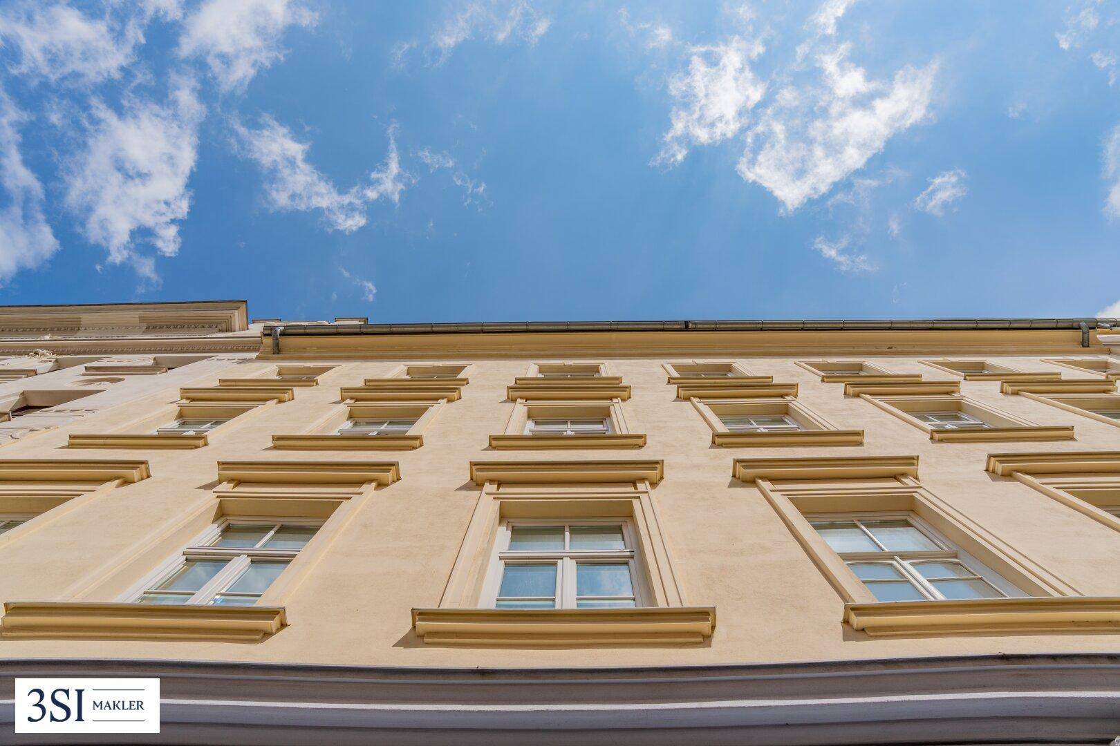 Blick nach oben auf die beige Fassade eines Altbaus mit vielen Fenstern unter blauem Himmel und Wolken.