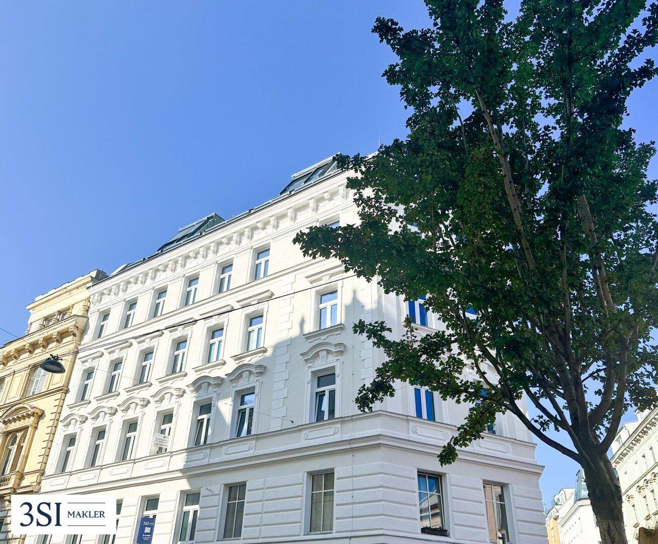 Seitenansicht eines eleganten Altbaus mit klassischer Architektur und einem großen Baum im Vordergrund.