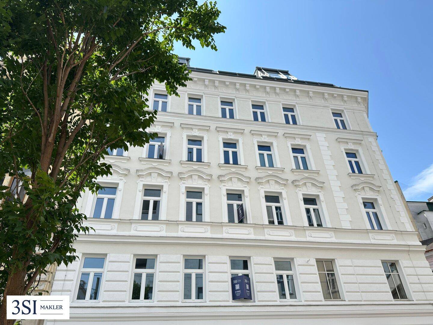 Helle, klassische Fassade eines Altbaus mit vielen Fenstern und grünen Bäumen im Vordergrund.