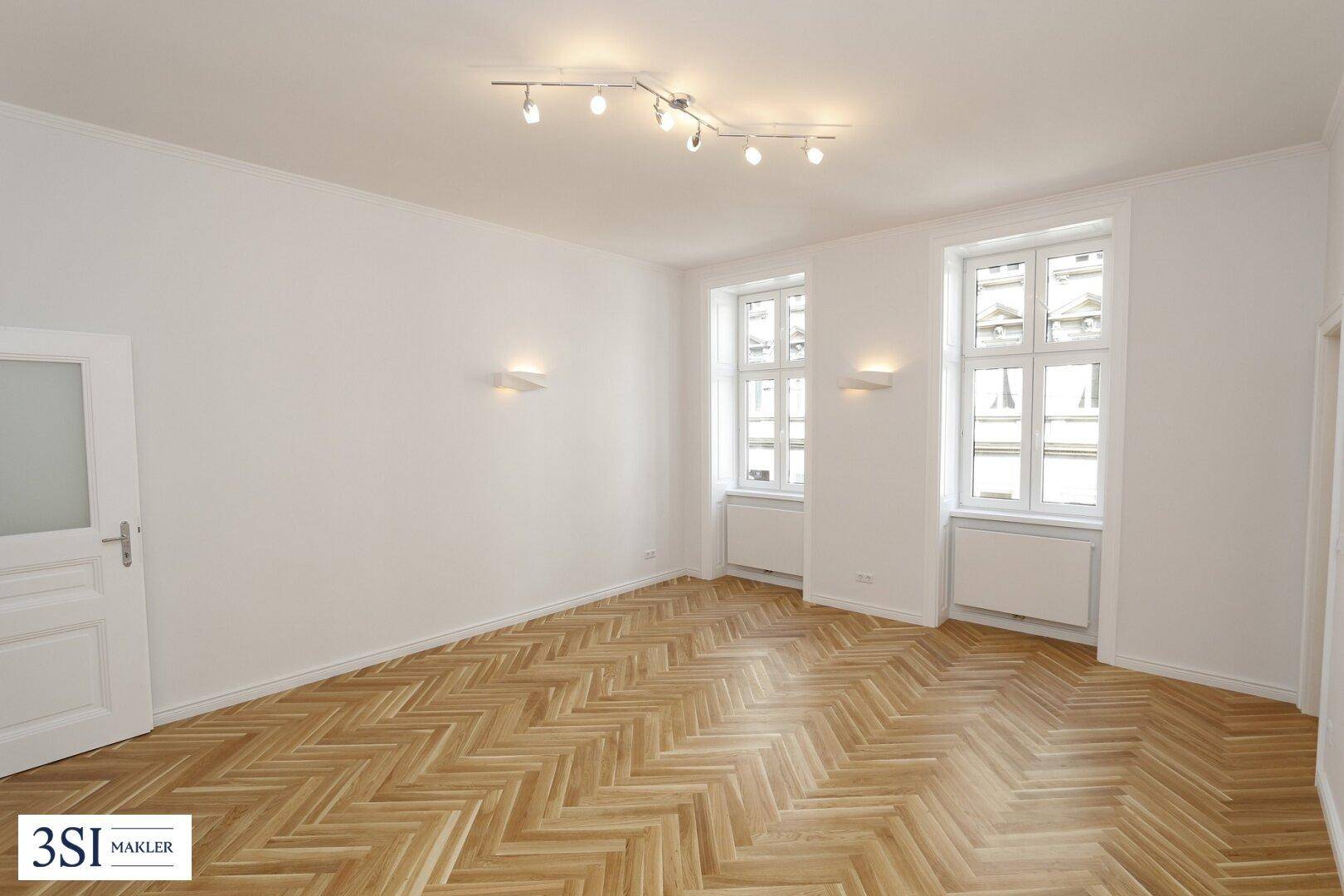 Geräumiges Zimmer mit hellem Fischgrätparkett, weißen Wänden und modernen Deckenstrahlern.