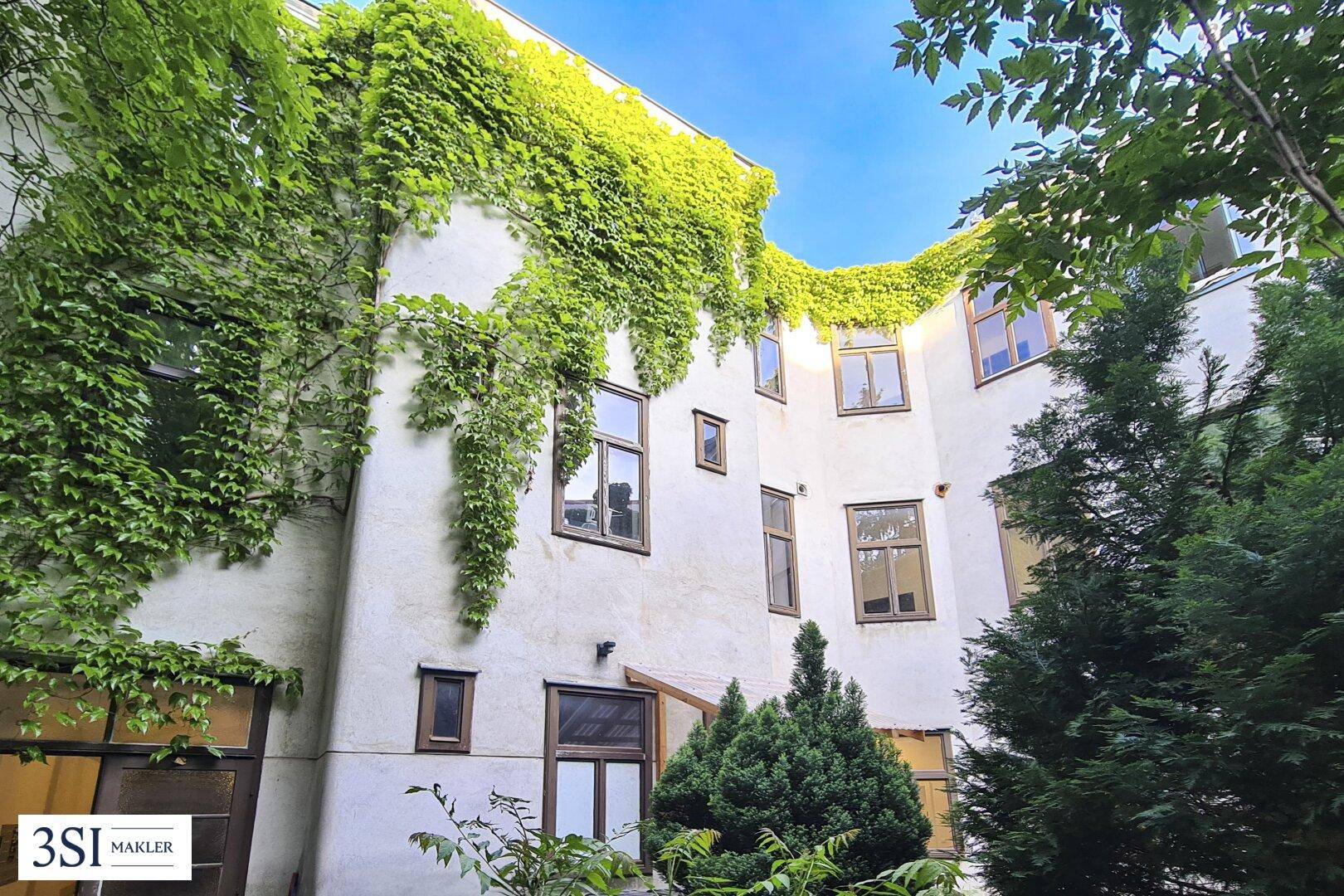 Grüne Efeuranken bedecken die Fassade eines Gebäudes mit vielen Fenstern, umgeben von Bäumen.