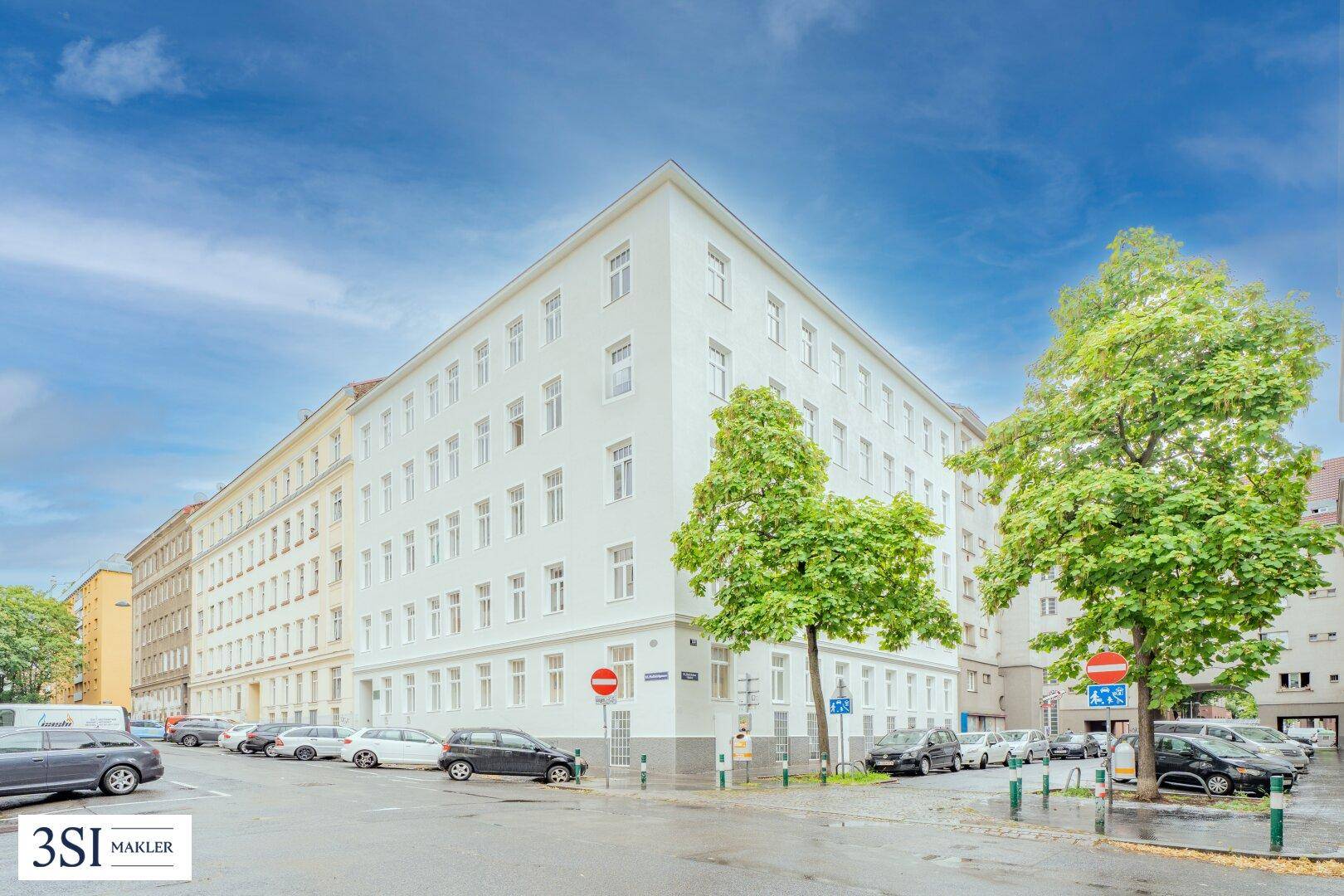 Weißes Eckgebäude mit vielen Fenstern an einer städtischen Straße mit parkenden Autos.