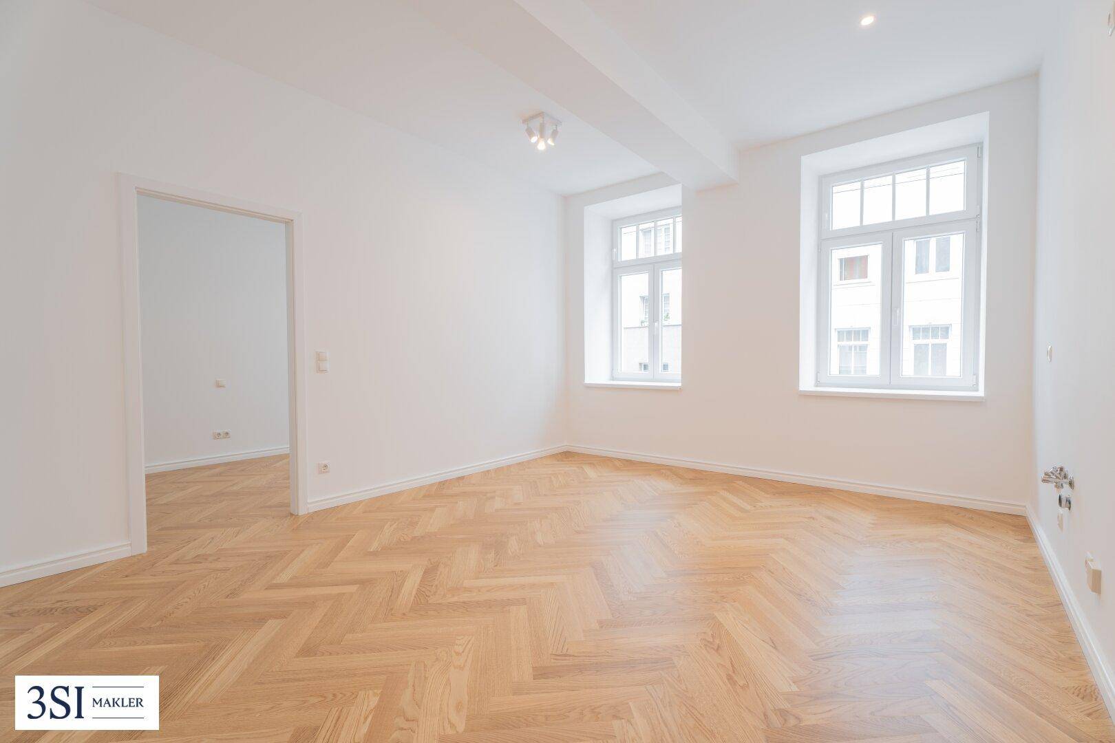 Geräumiges, leeres Zimmer mit Fischgrätparkett und zwei großen Fenstern, bereit zur Einrichtung.