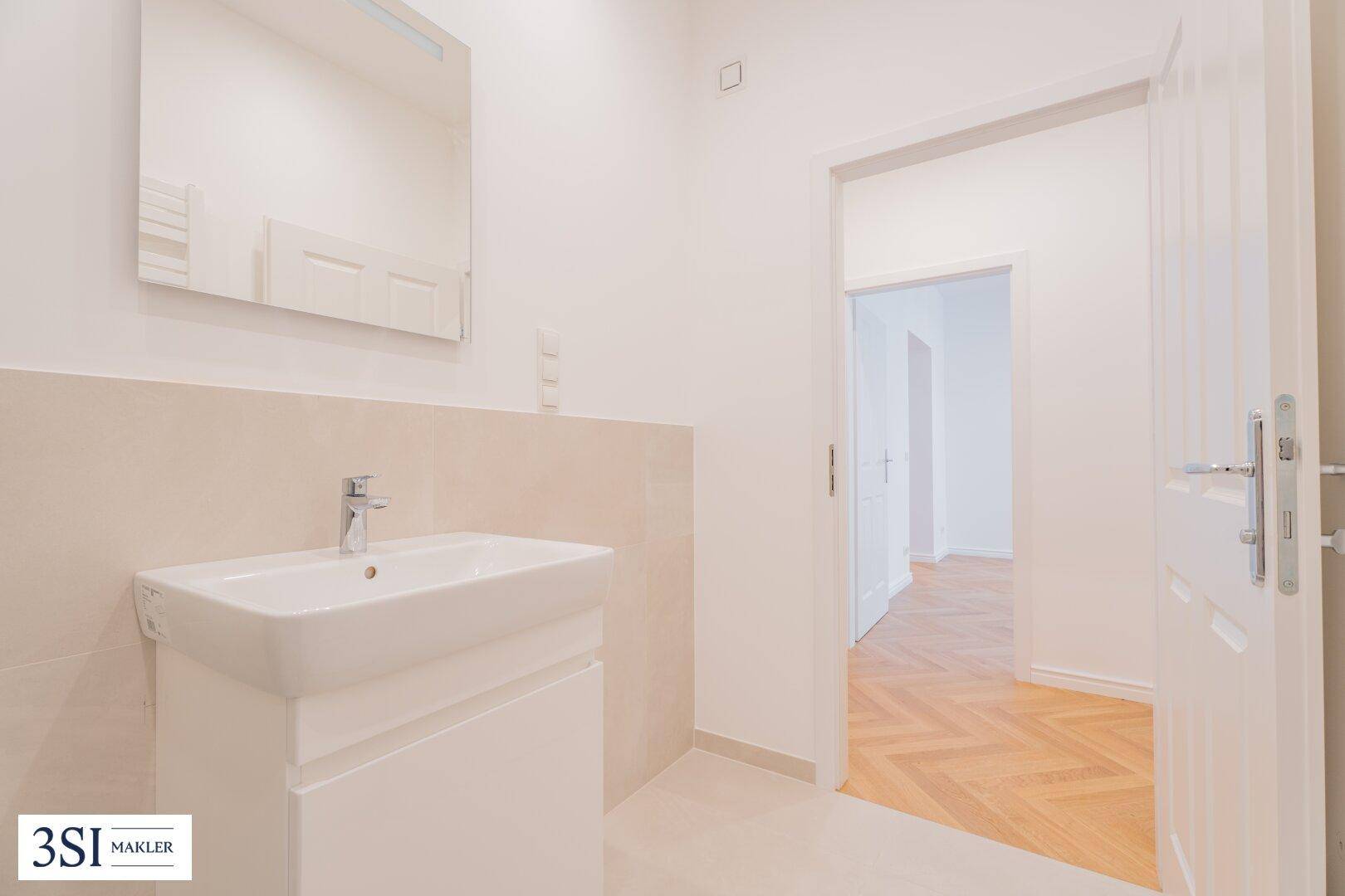 Helles Badezimmer mit Waschbecken, Spiegel und Zugang zum Flur.