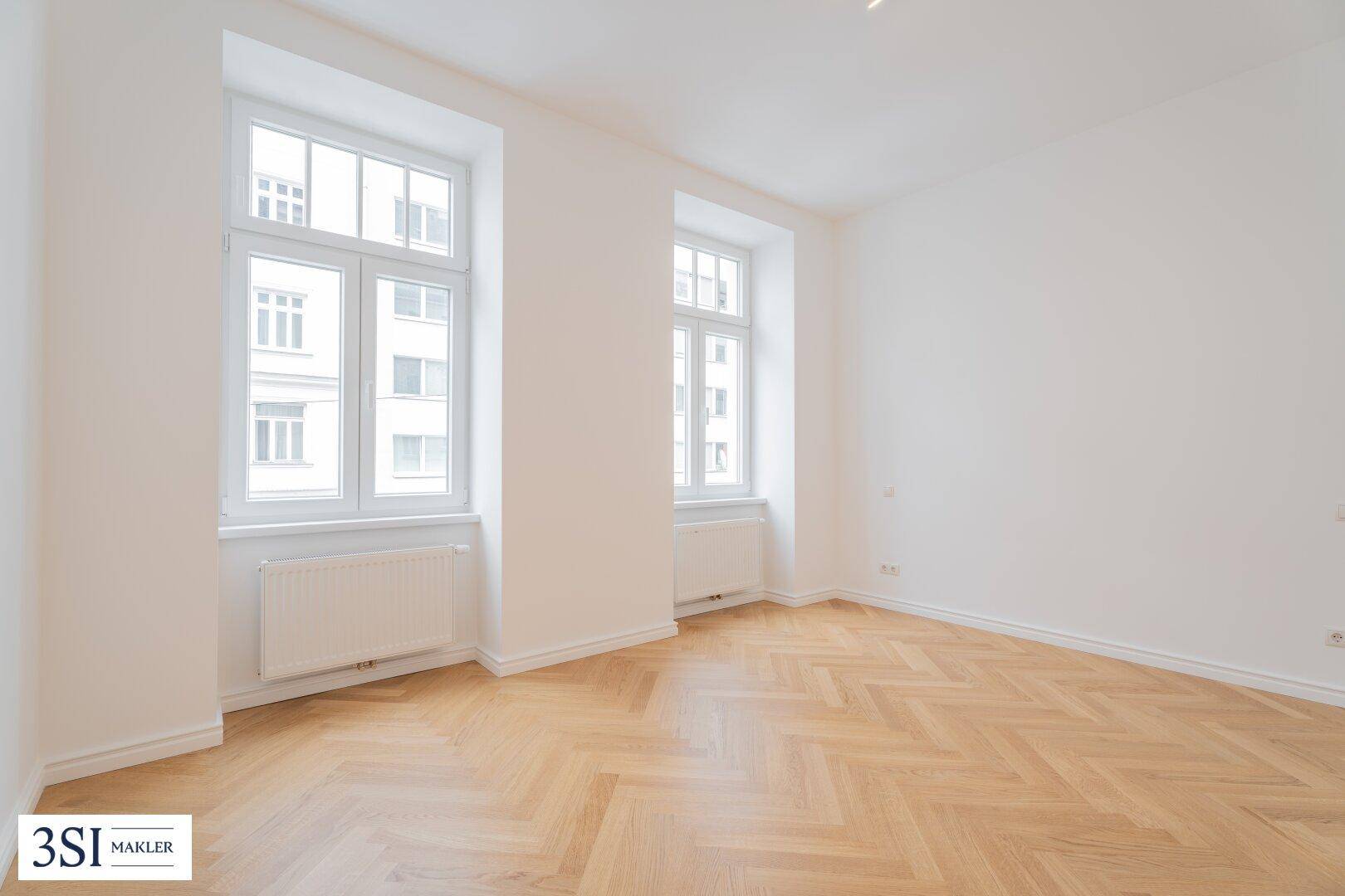 Großes, helles Zimmer mit Fischgrätparkett und zwei hohen Fenstern.