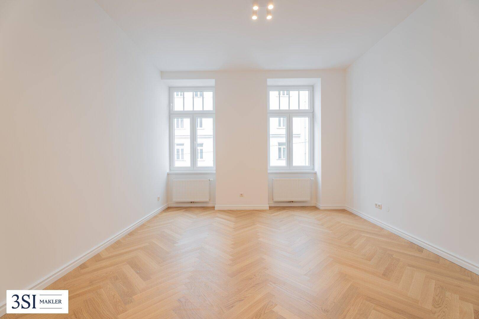Geräumiges, helles Zimmer mit Fischgrätparkett und zwei großen Fenstern.