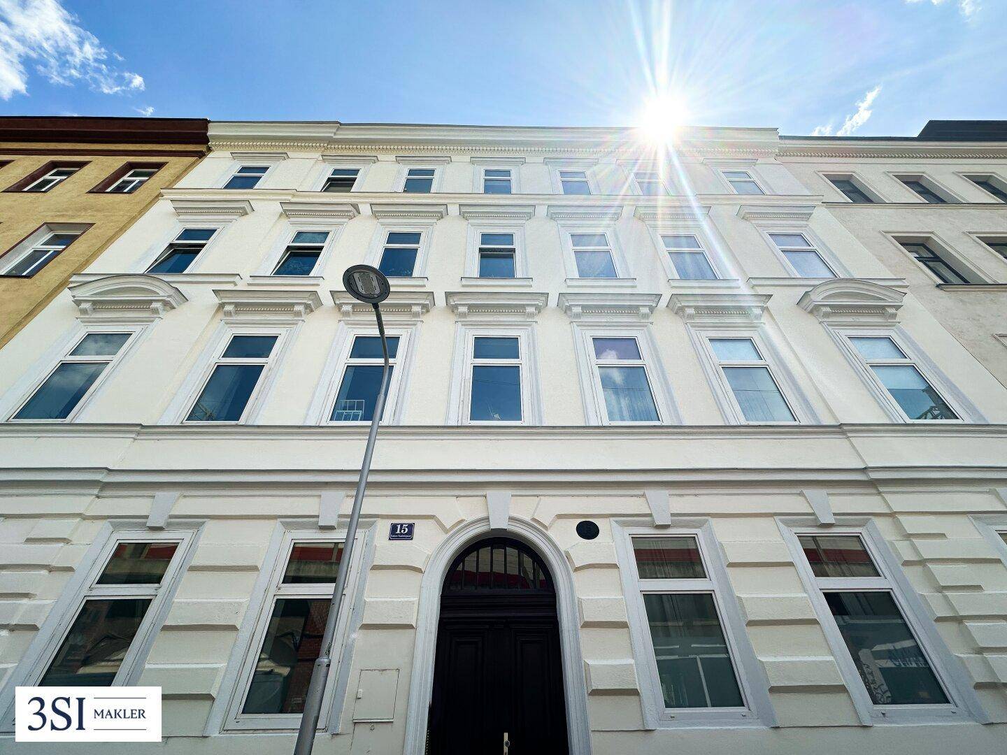 Helle Fassade eines klassischen Wohngebäudes mit vielen Fenstern unter strahlendem Sonnenschein.