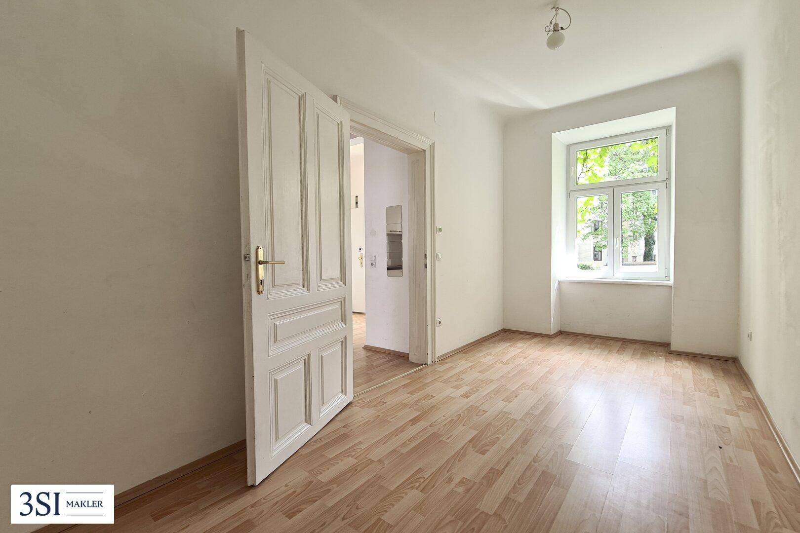 Leerer Raum mit Holzboden, weiß gestrichenen Wänden und einem Fenster zum Innenhof.
