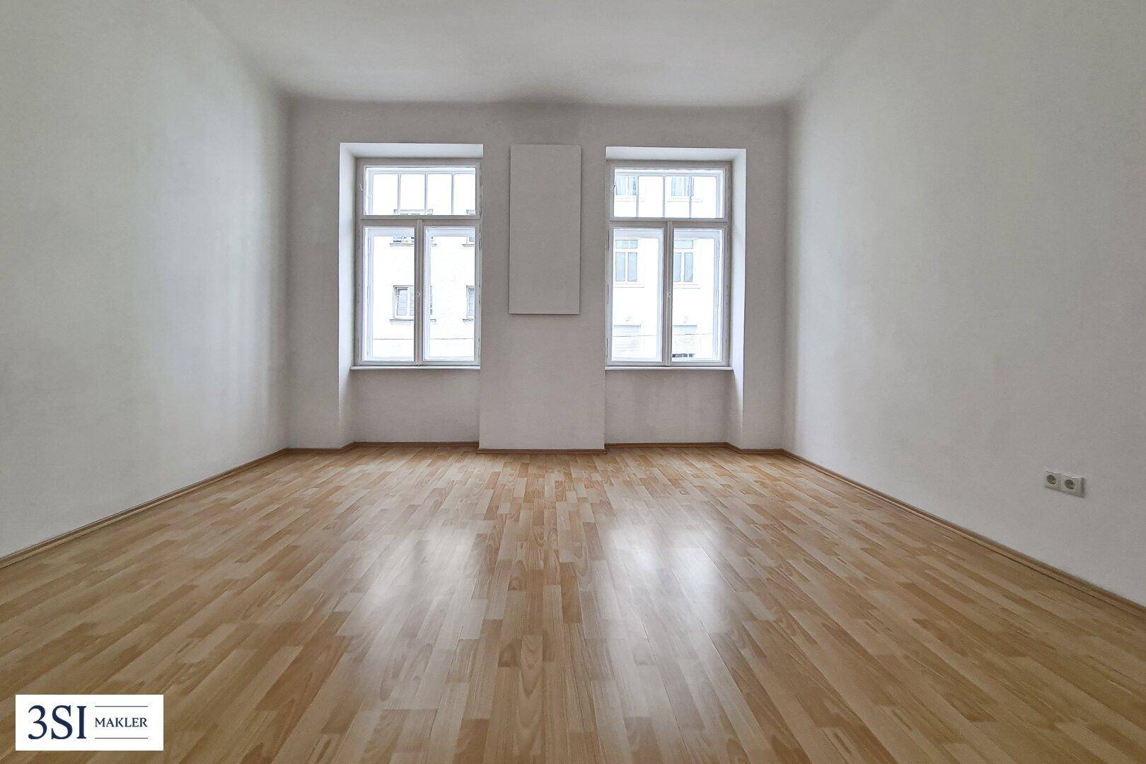 Heller, leerer Raum mit Holzboden und zwei großen Fenstern.