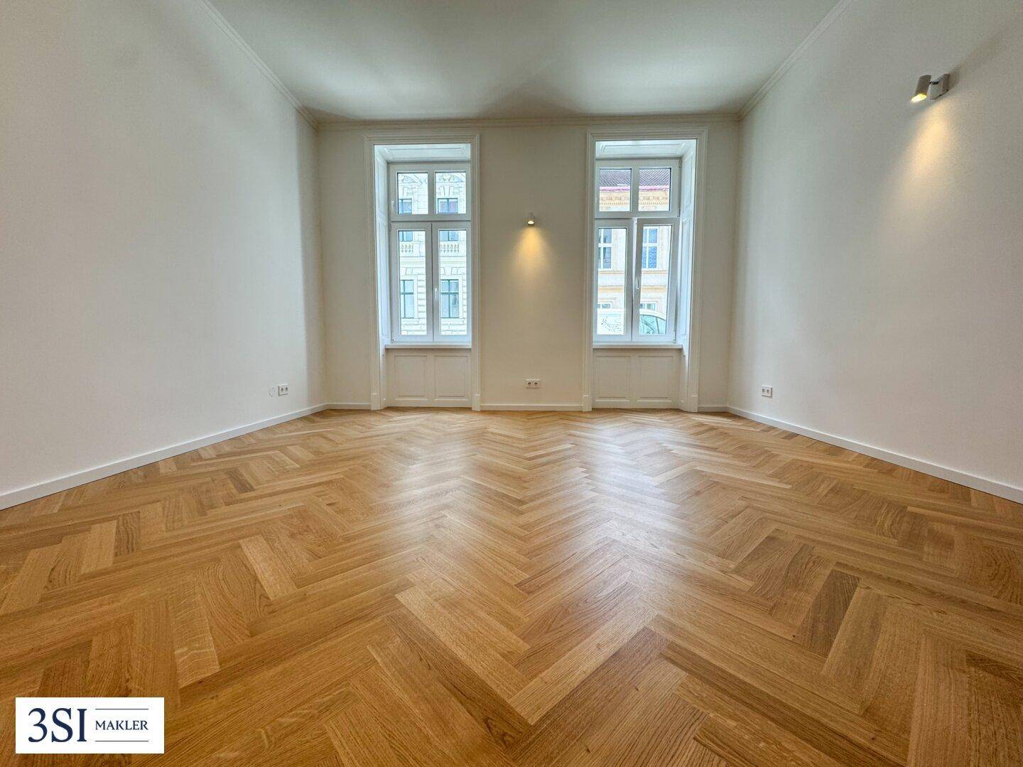 Heller, leerer Raum mit zwei großen Fenstern und elegantem Fischgrätparkettboden.