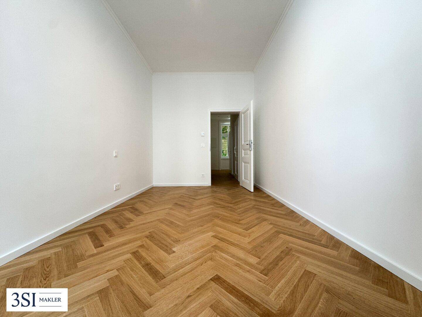 Leerer Raum mit klassischem Fischgrätparkett und weiß gestrichenen Wänden, mit Blick in den Flur.
