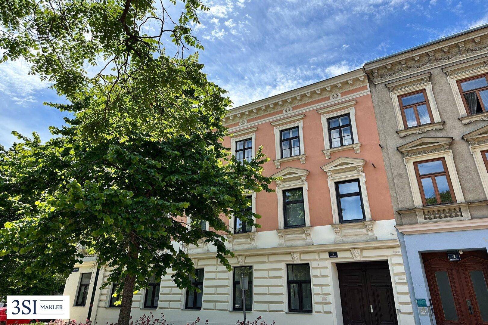 Historische Fassade eines Gebäudes mit rosa Putz und weißen Verzierungen, umgeben von grünen Bäumen.