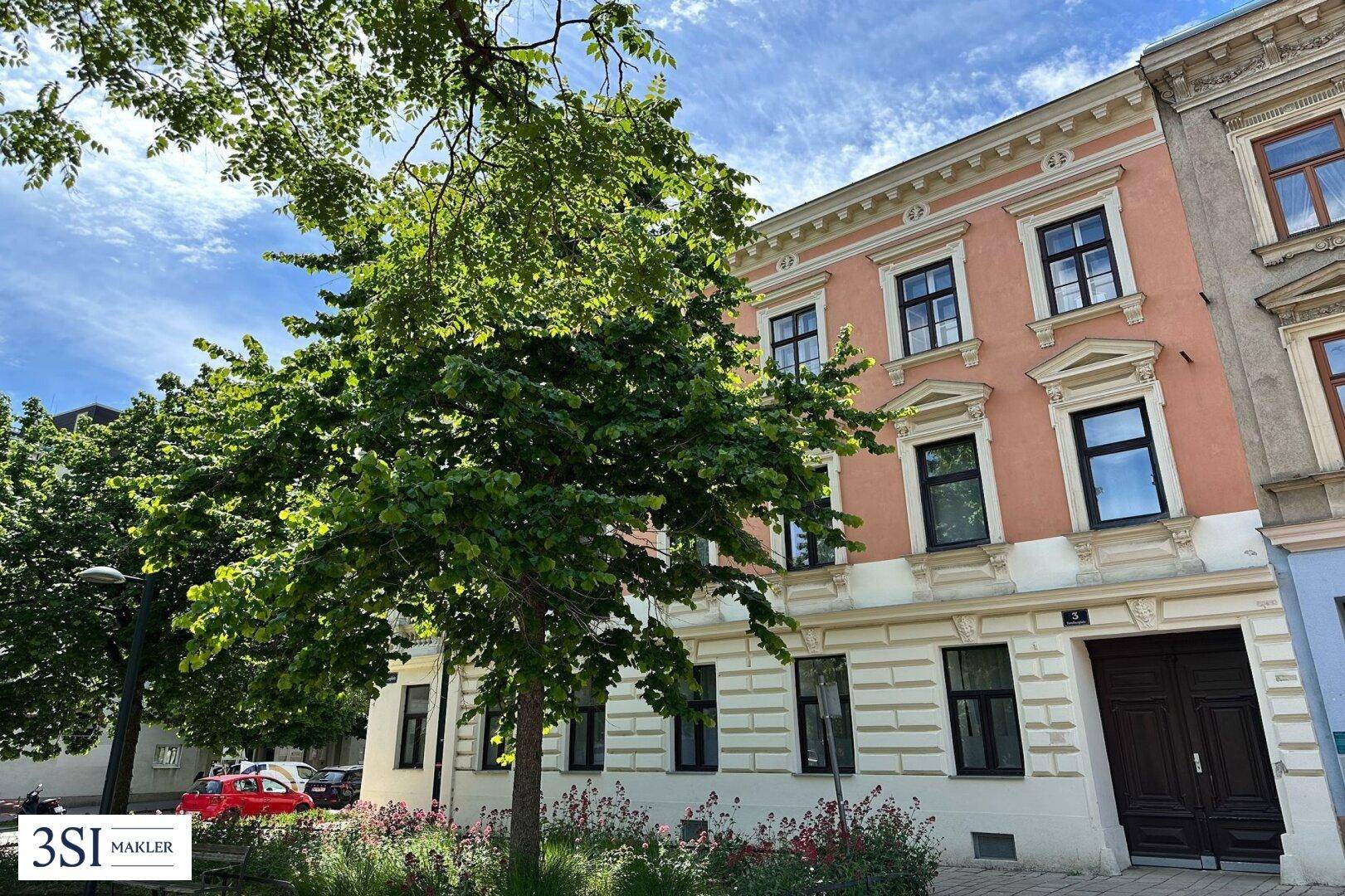 Historische Fassade eines Wohngebäudes mit dekorativen Elementen und grünen Bäumen davor.