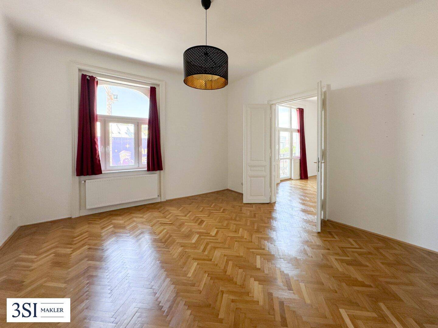 Heller Raum mit Fischgrätparkettboden, weißen Wänden und einem Fenster mit roten Vorhängen.