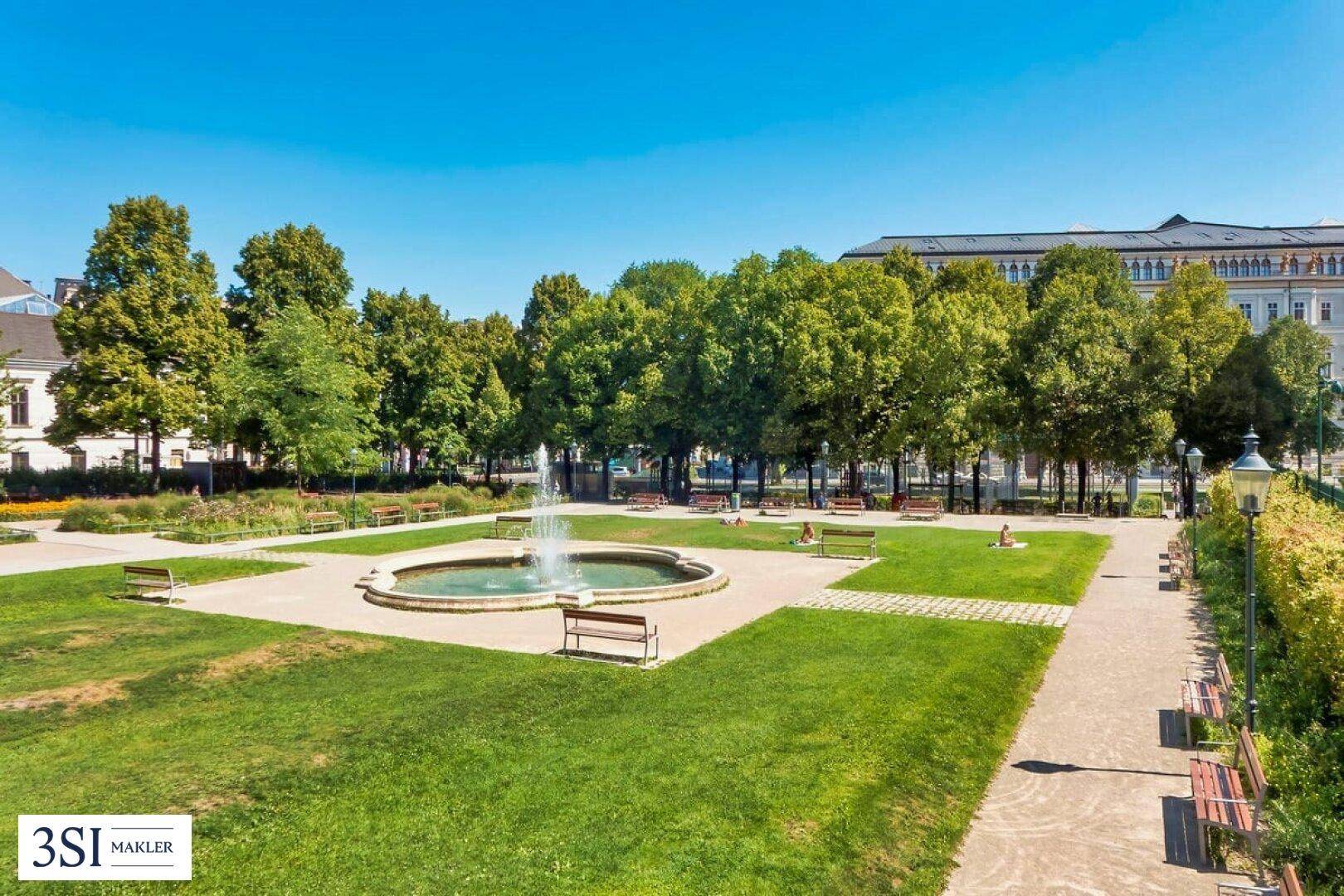 Grüne Parkanlage mit Springbrunnen und Sitzbänken unter blauem Himmel.